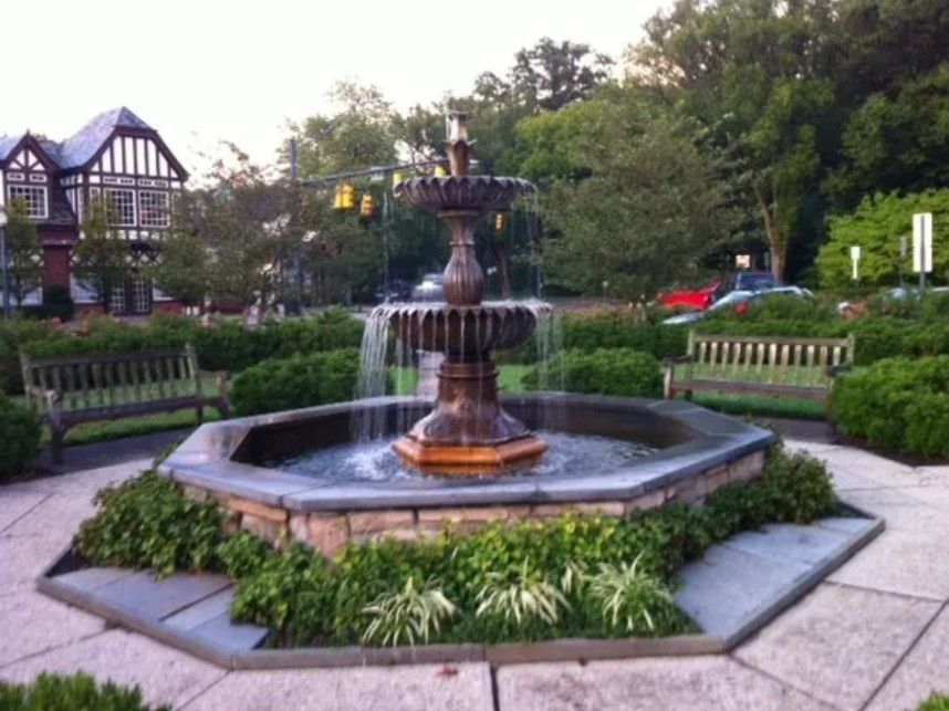 A multi-tiered metal fountain centered in a stone plaza, surrounded by benches, greenery, and a Tudor-style building.