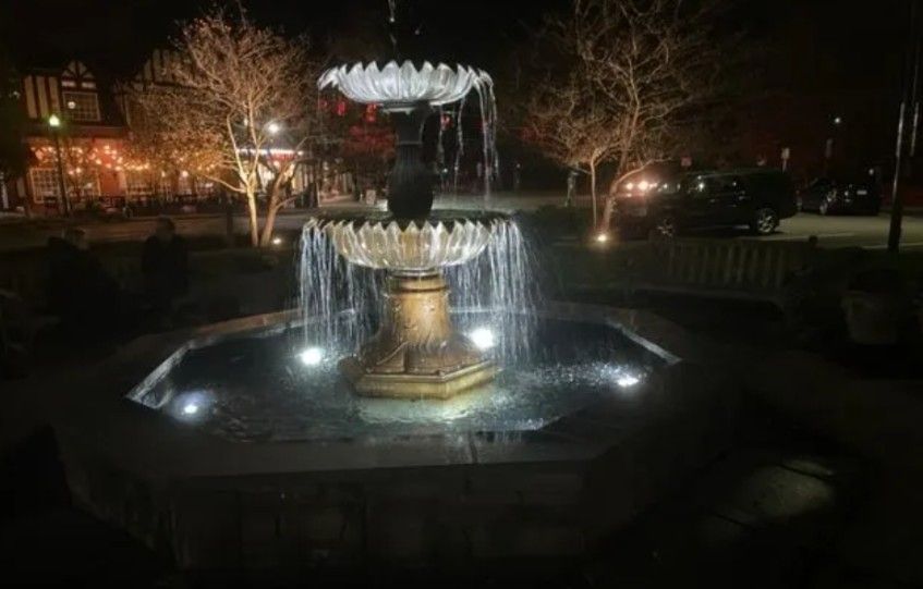 A multi-tiered stone fountain illuminated at night, surrounded by trees and distant building lights.