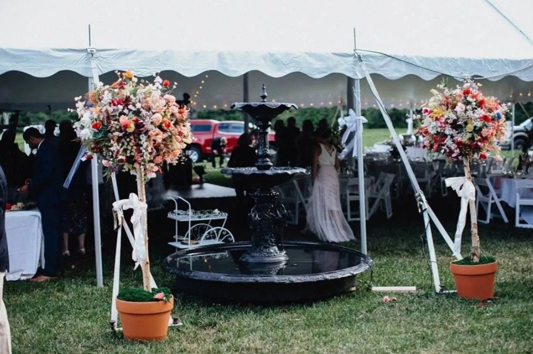 A tiered fountain sits between two large potted floral arrangements under a white event tent at an outdoor gathering.