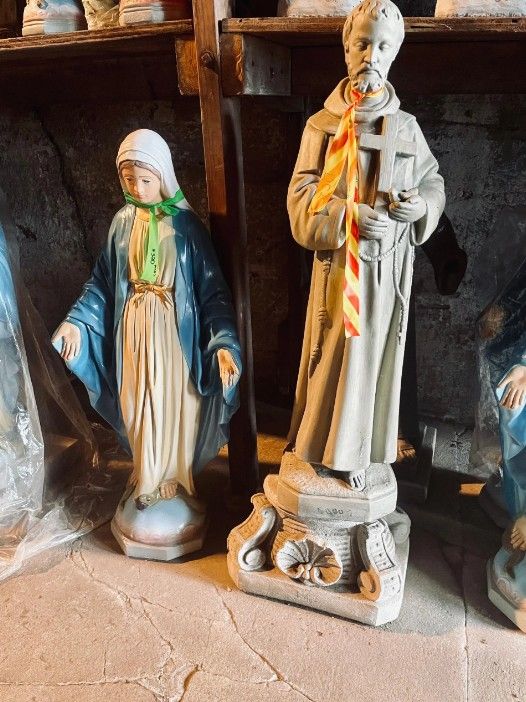 Two religious statues stand on a shelf, one of a draped figure and one holding a cross, adorned with a striped neck tie.