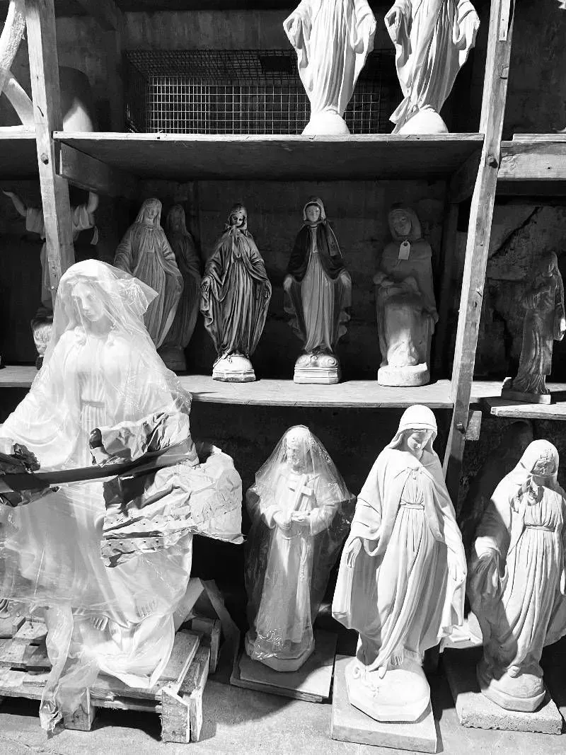 A workshop shelf holds rows of various white, carved religious statues of figures in robes and veils.