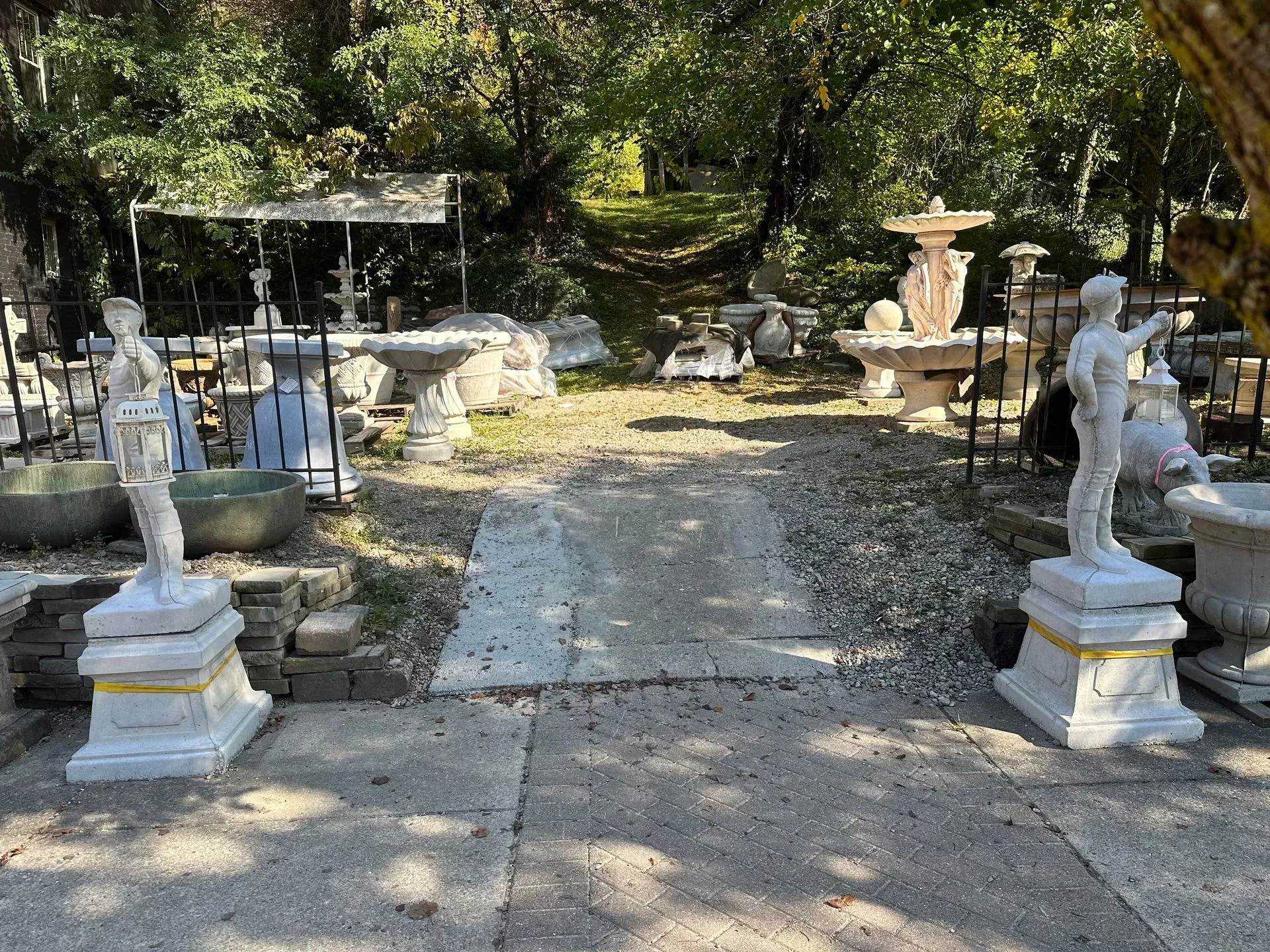 Two white statues flank a gravel path leading to a garden area filled with various concrete planters and fountains.