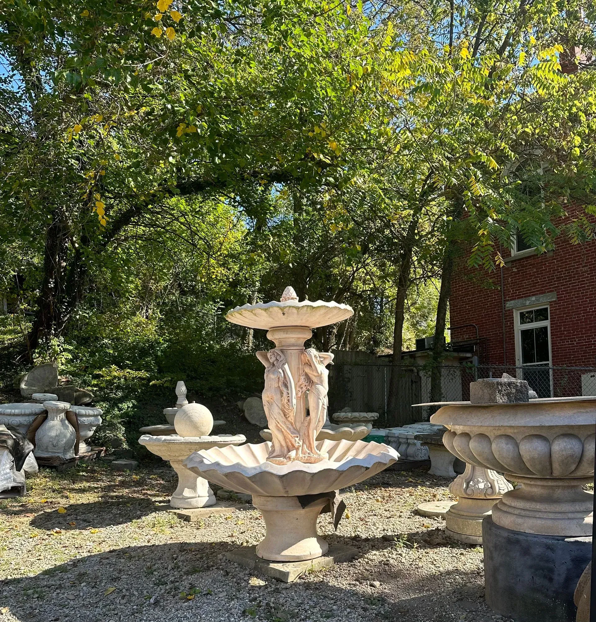 Several ornate stone fountains sit in a gravel yard, surrounded by lush green trees and a red brick building.