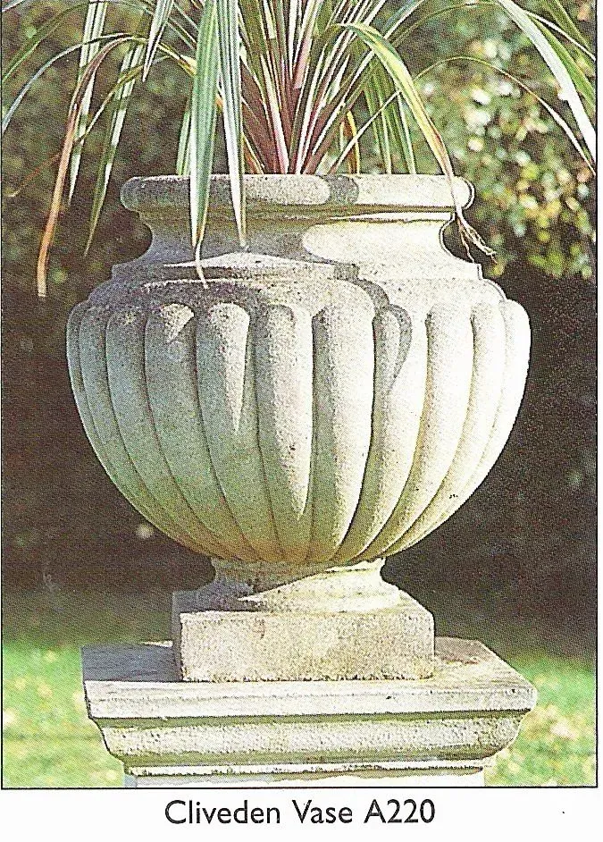 A fluted, stone-colored garden urn with a plant inside, set on a square pedestal against a blurred outdoor background.