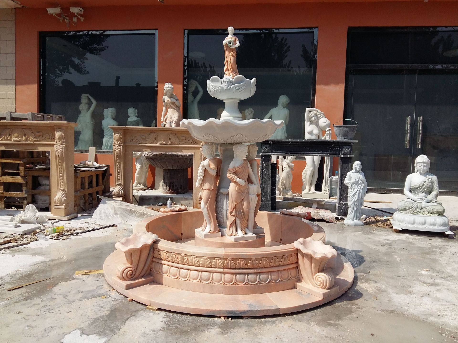 A tiered stone fountain surrounded by various marble statues, carvings, and decor in front of an orange-walled building.