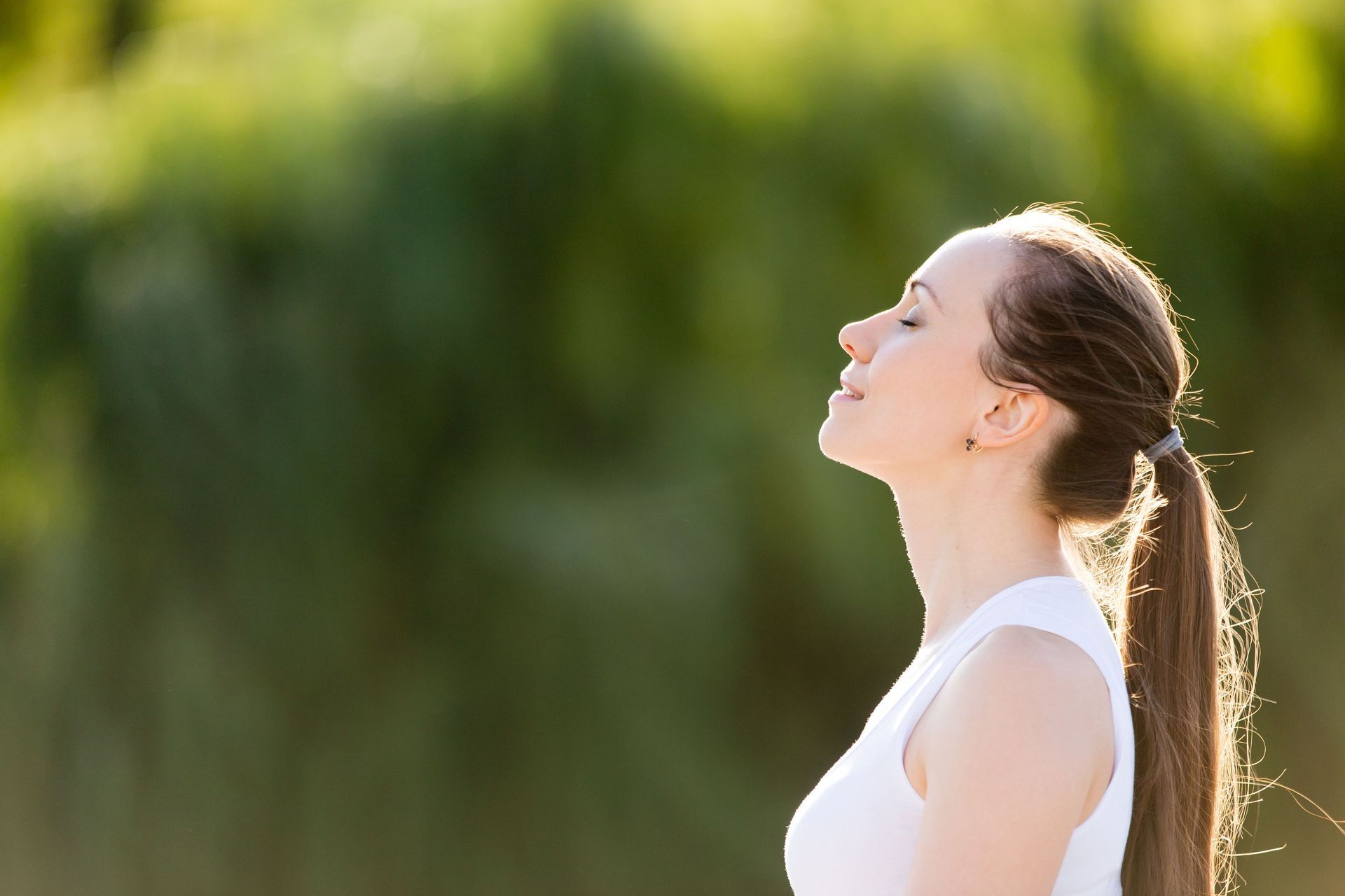 Woman with eyes closed, tilting head back, breathing outdoors with sunlight.