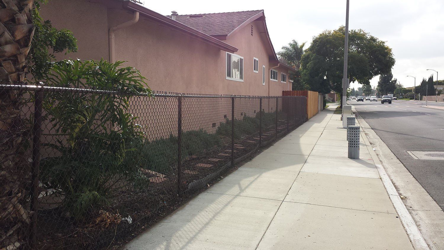 Sidewalk alongside a residential building with a chain-link fence and street traffic.