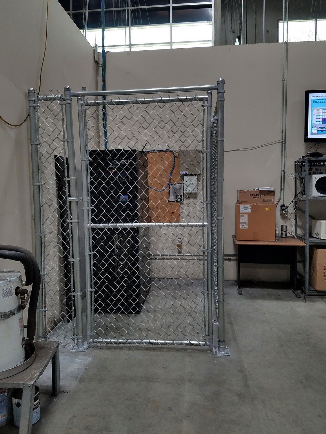 A chain-link fenced enclosure with a locked gate, containing equipment, in an industrial setting.