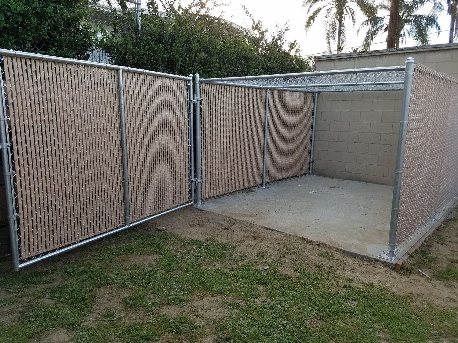 A small fenced-in area, open gate, tan and silver fencing, in a grassy yard.