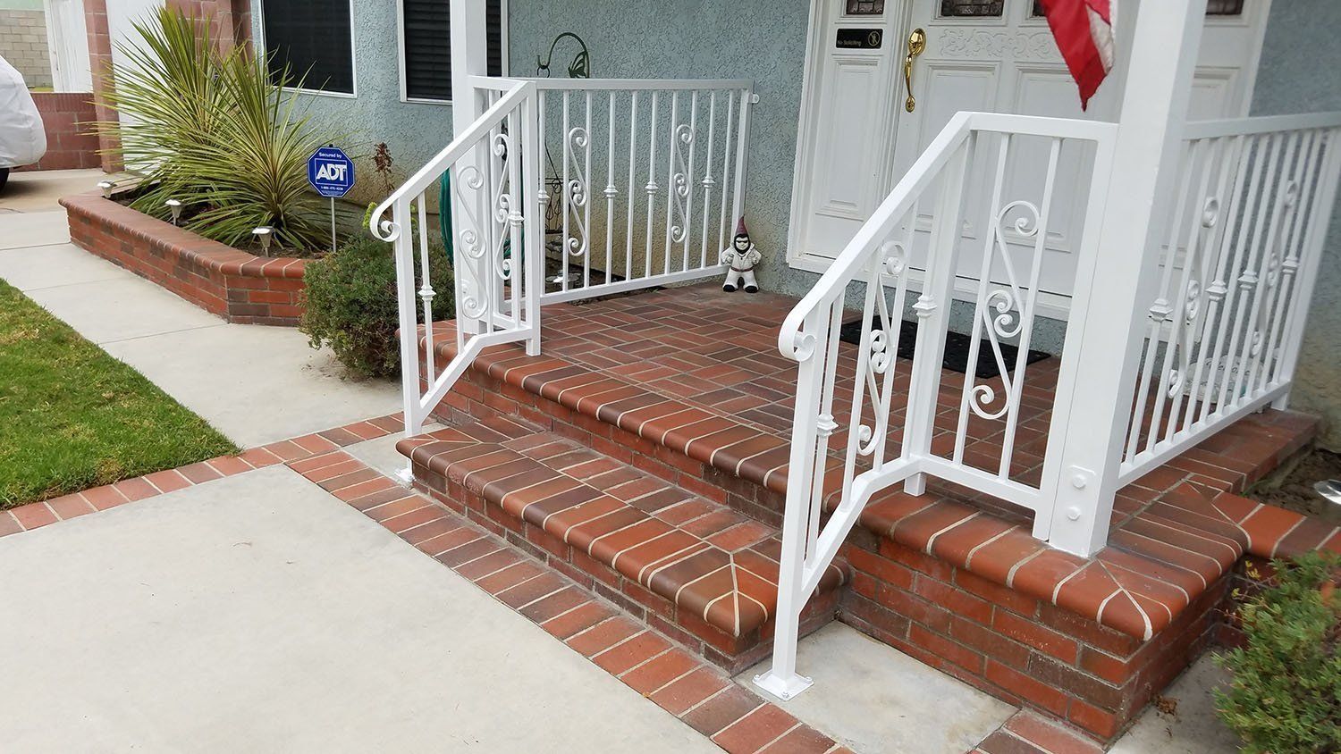 White railing on brick steps leading to a house with a small porch and garden bed.