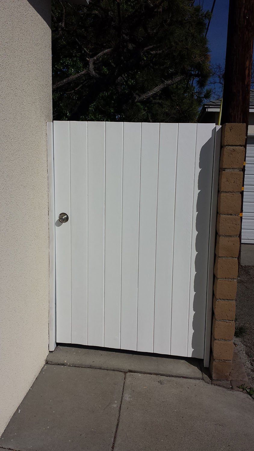 White gate on a concrete path, between a stucco wall and brick column, leading to a backyard.