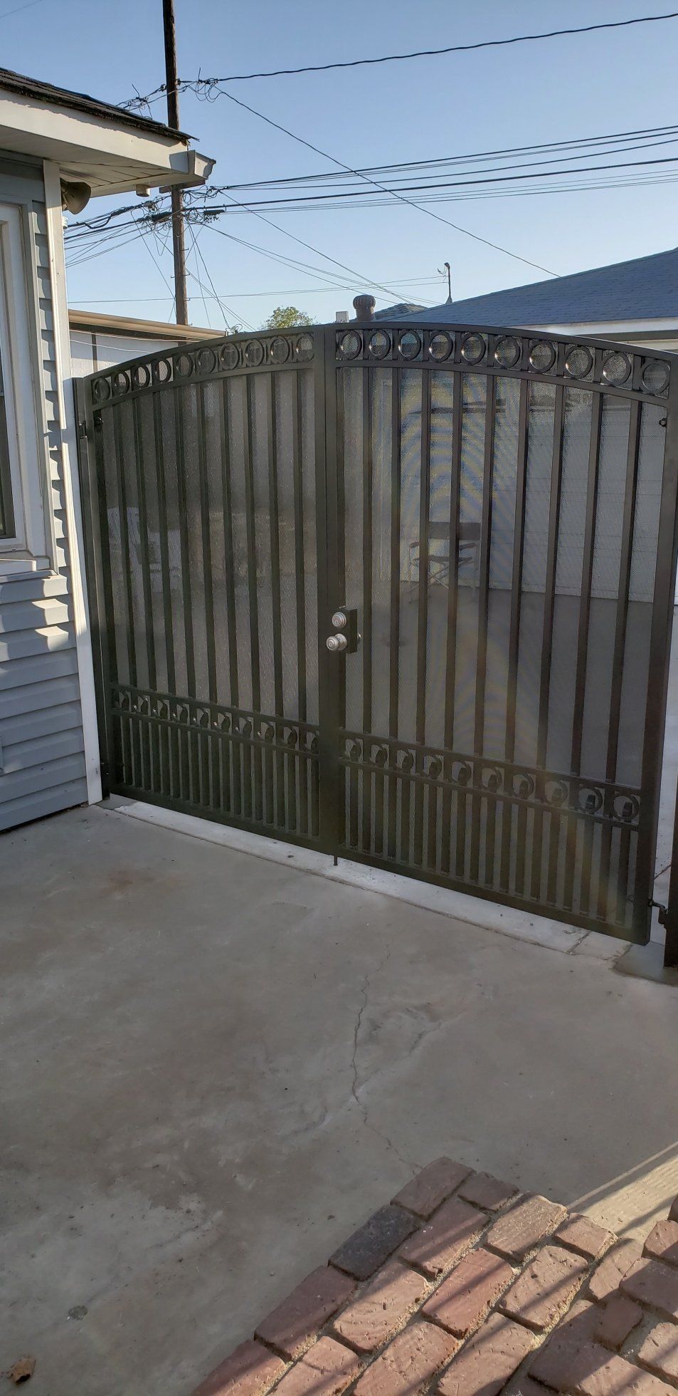 Metal gate in front of a gray house. Brick pavers are visible on the ground.
