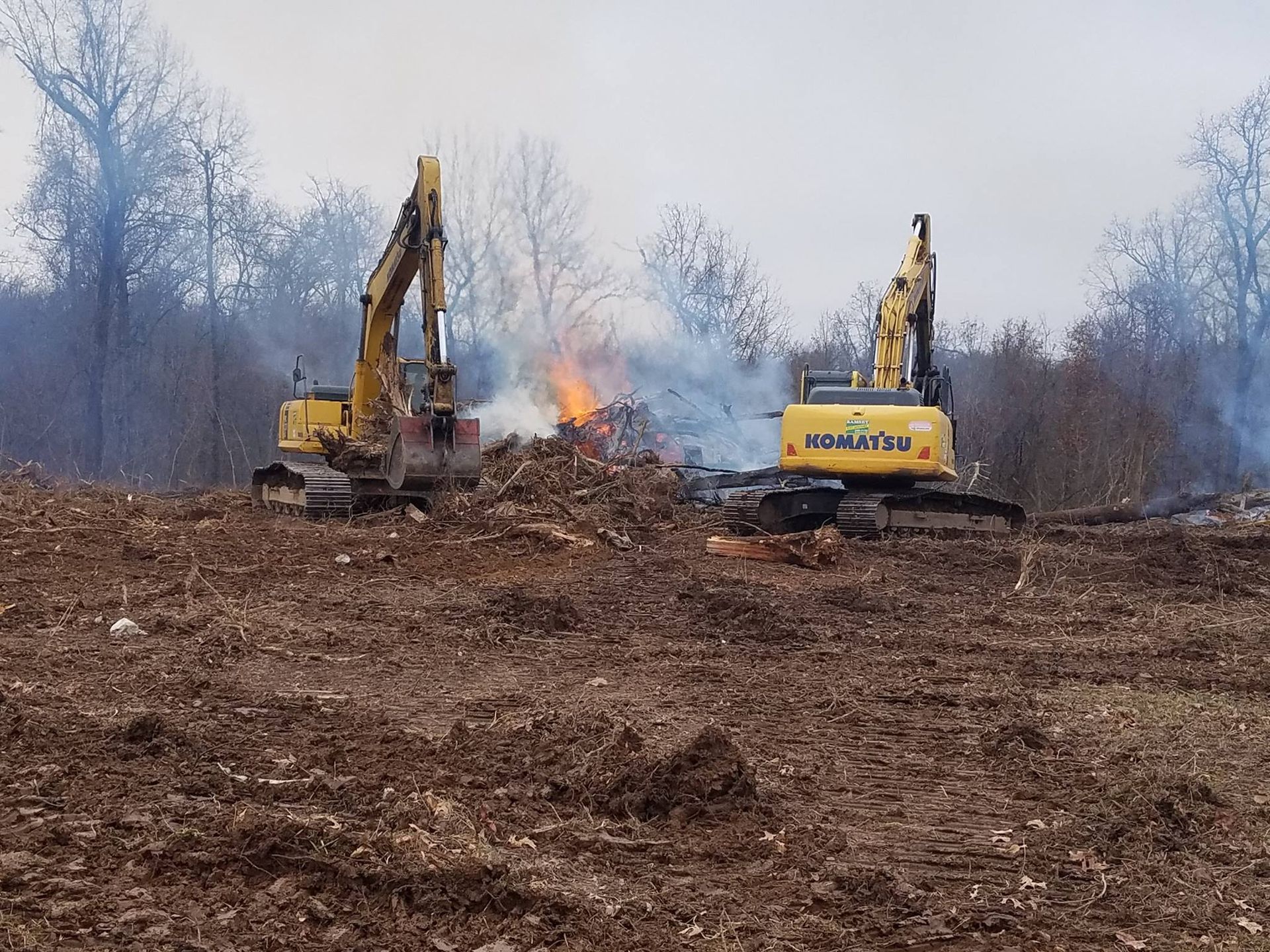Two bulldozers are working in a field with a fire in the background.