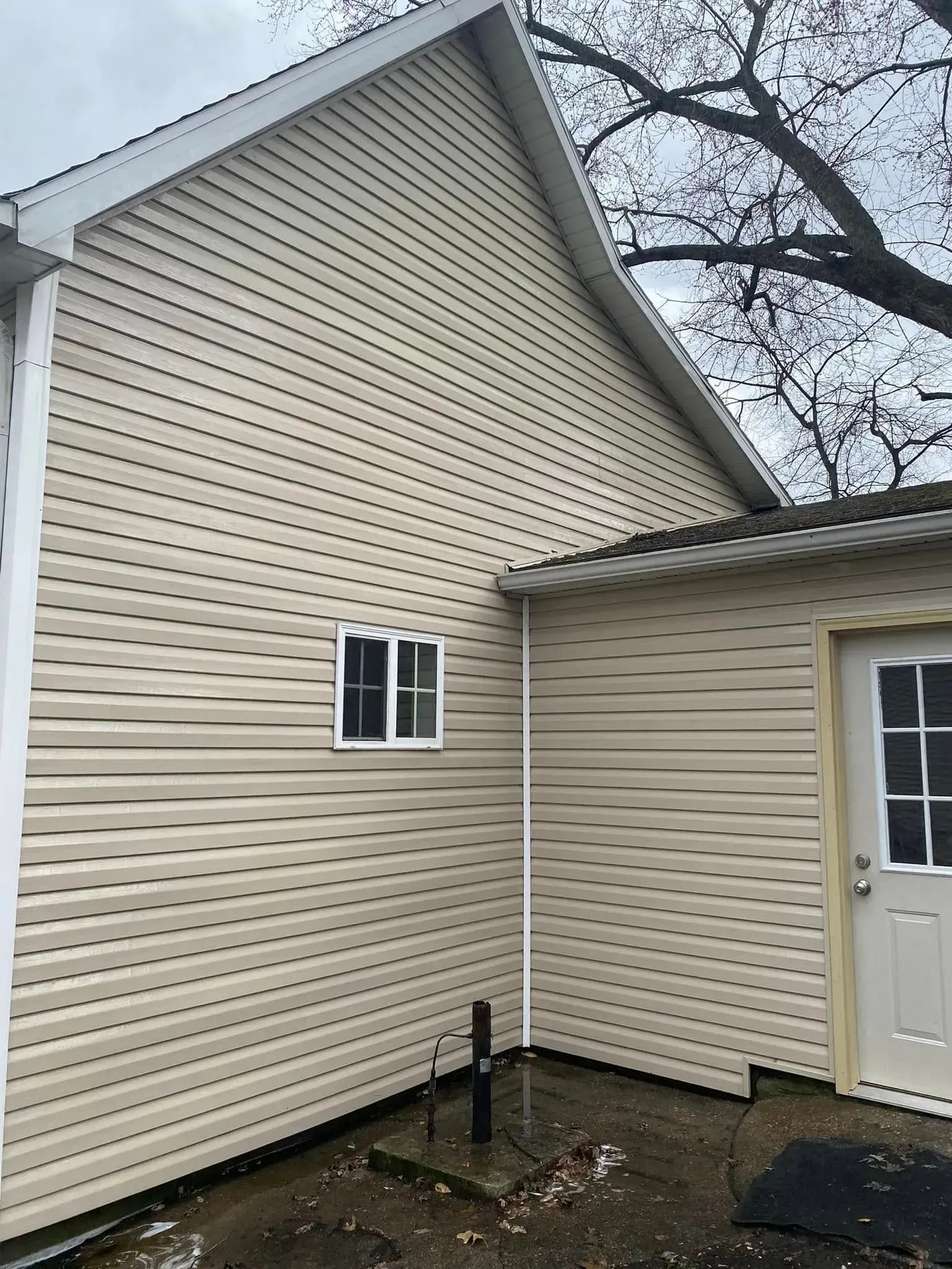 Beige house siding and roof corner with a small window and side entrance door under bare tree branches.