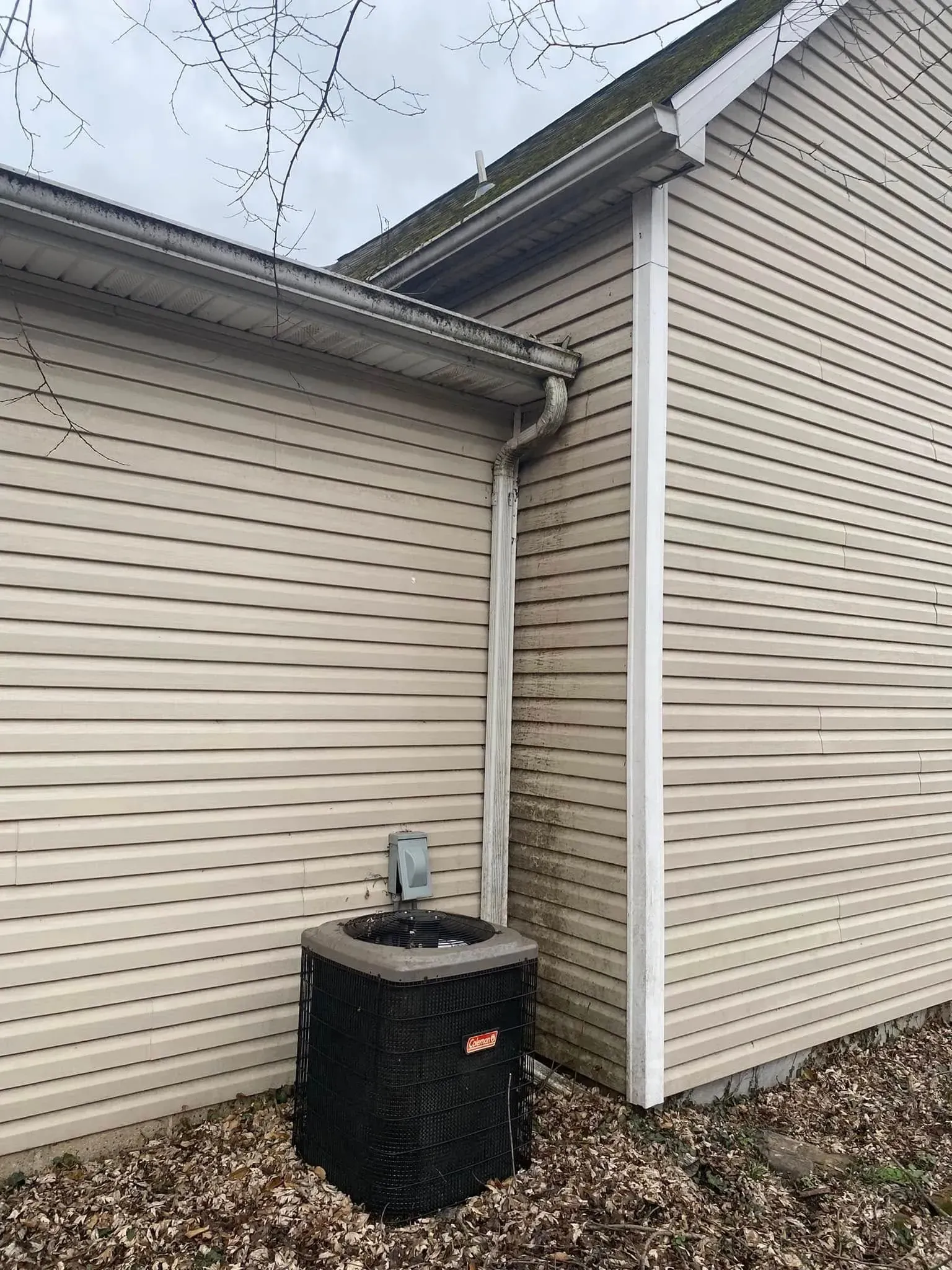 Outdoor house siding corner with beige vinyl walls and a black air-conditioning unit on gravel.
