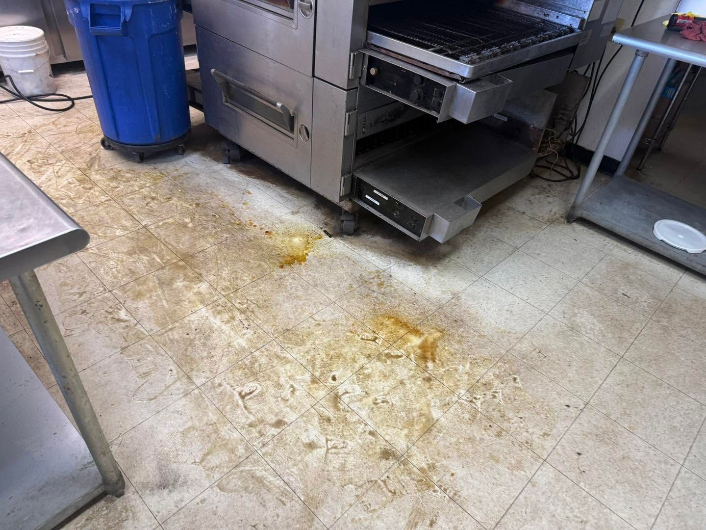 Dirty commercial kitchen floor beside stainless steel equipment and a blue trash bin