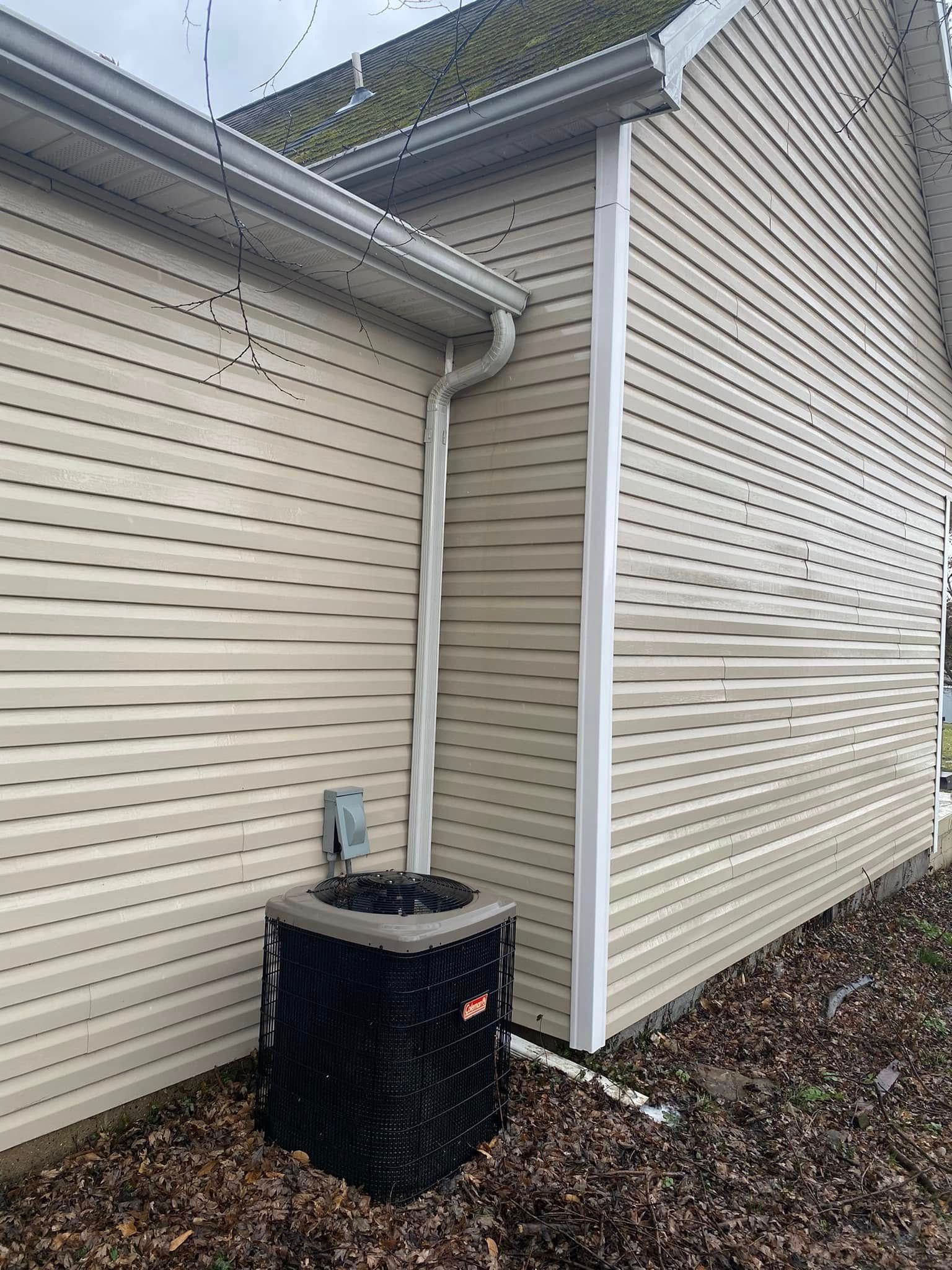 Outdoor HVAC unit beside beige house siding and downspout under a covered eave.