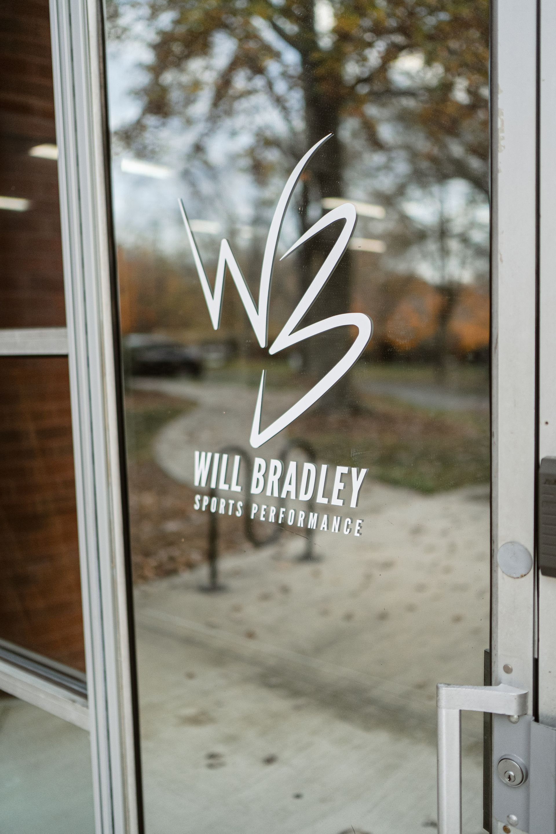 Glass door with white “Will Bradley” salon logo and lettering, reflecting an outdoor sidewalk and trees