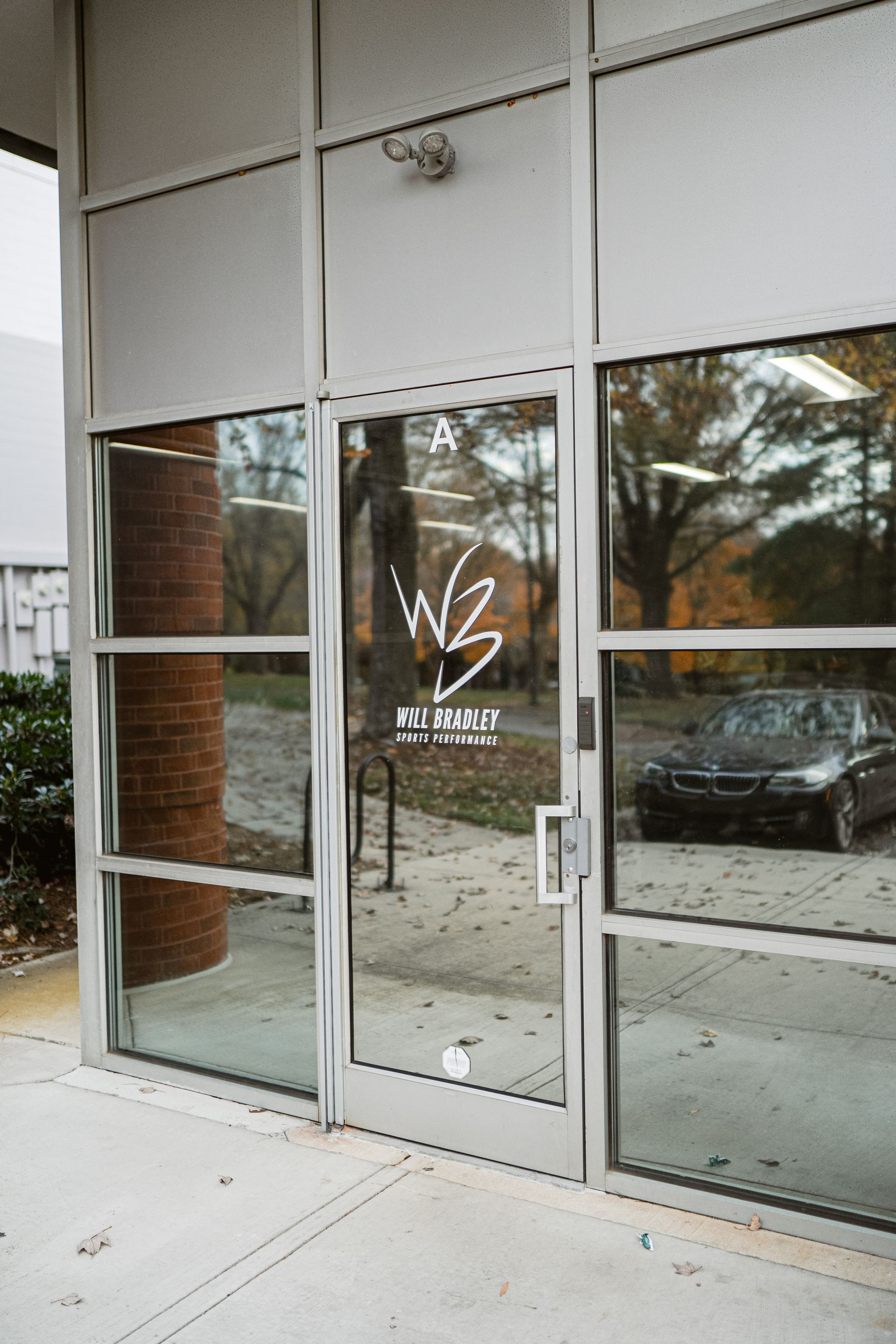 Glass storefront entrance with white-framed doors and windows reflecting trees and parked cars.