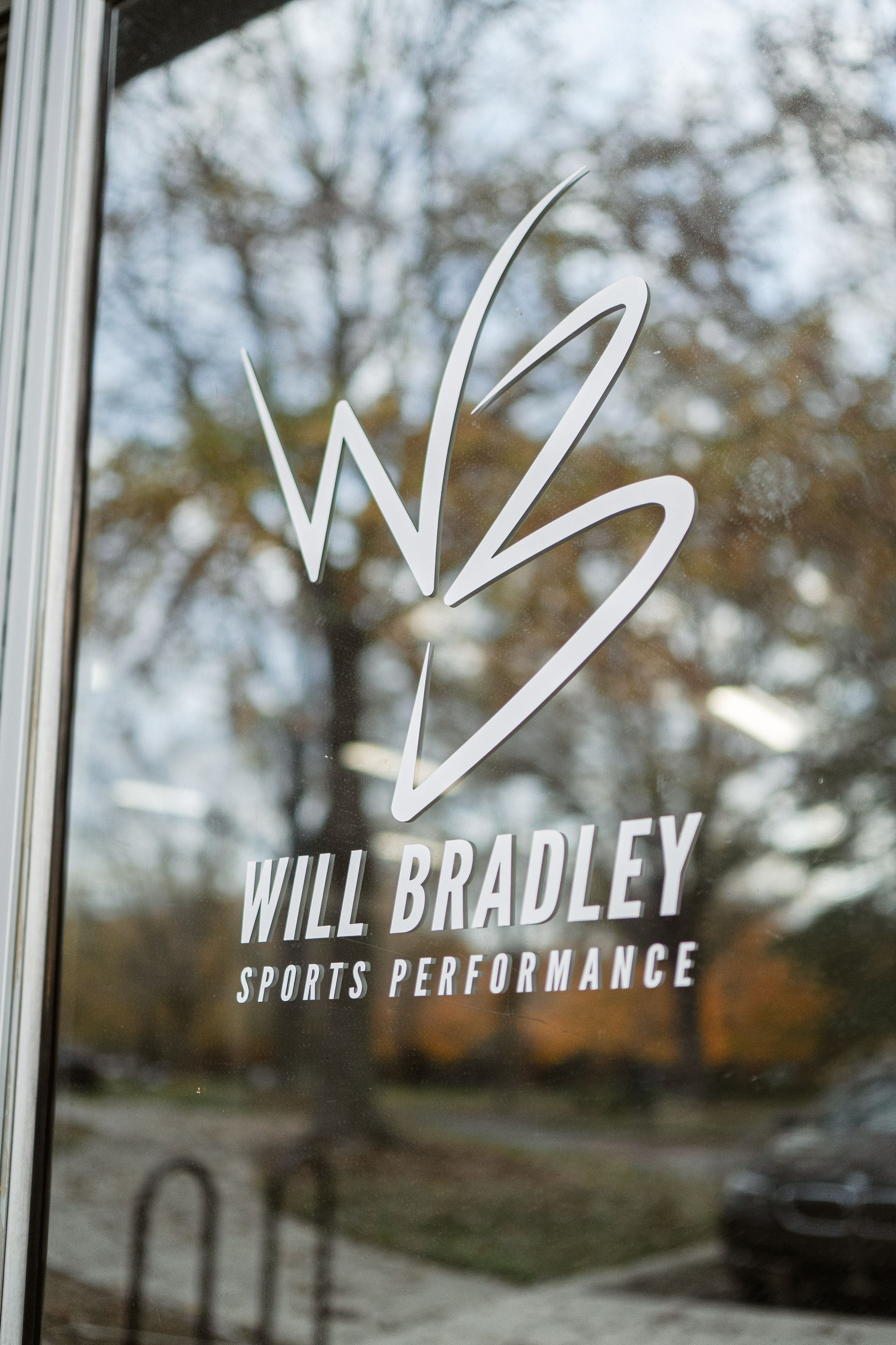 Will Bradley Sports Performance logo on glass with blurred trees and parked cars in the background