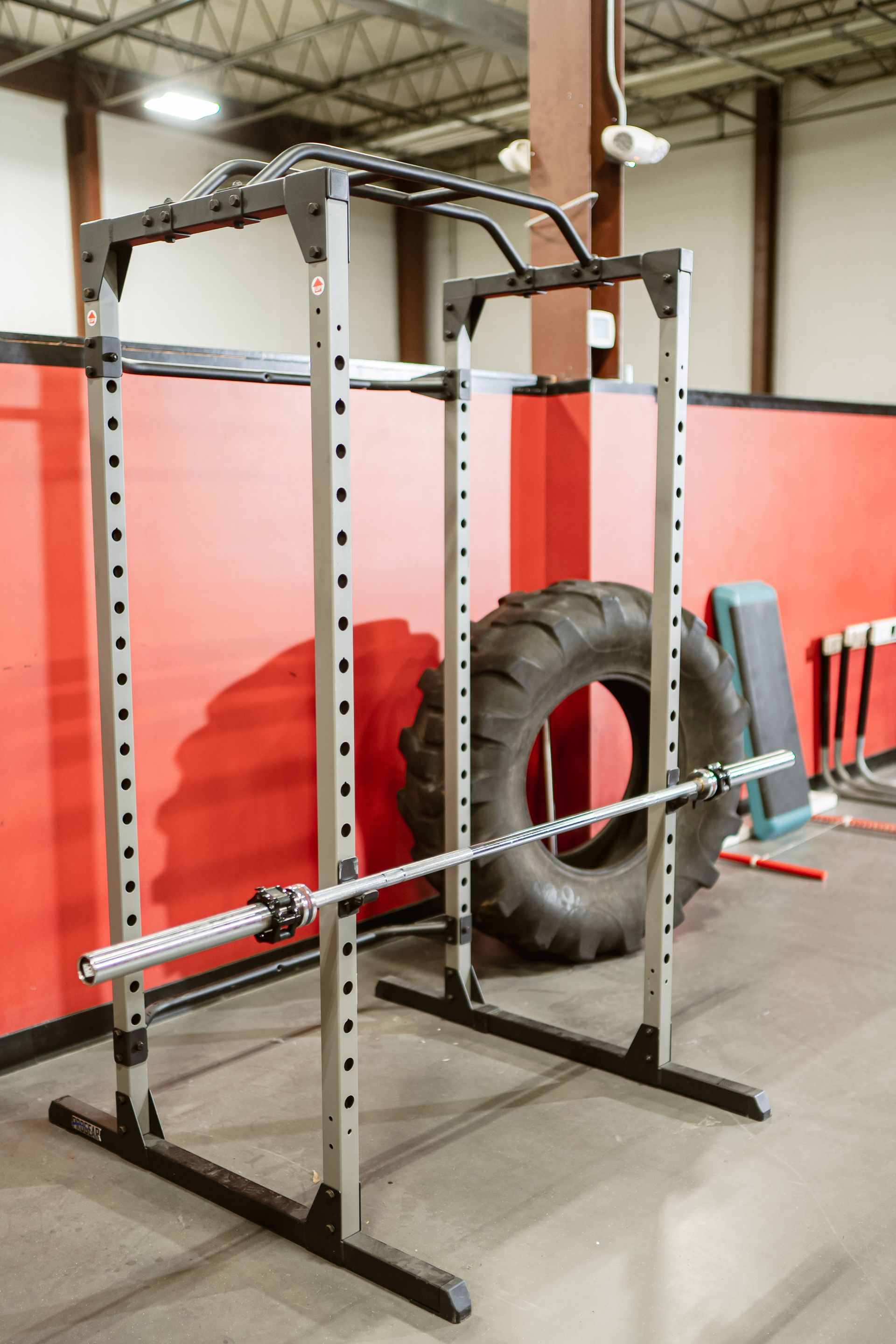 Industrial squat rack with a barbell, large tire, and red wall panels in a gym.