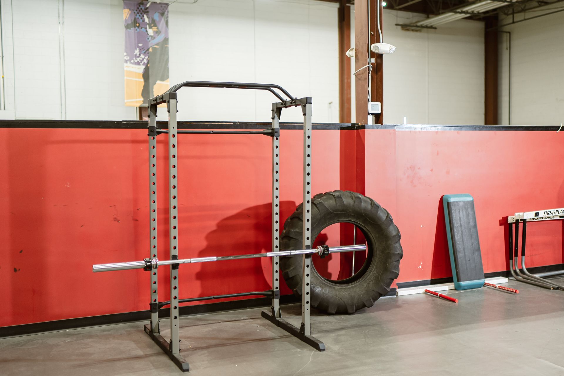 Gym rack with pull-up bar, tire, and workout equipment against a red wall in a garage gym.