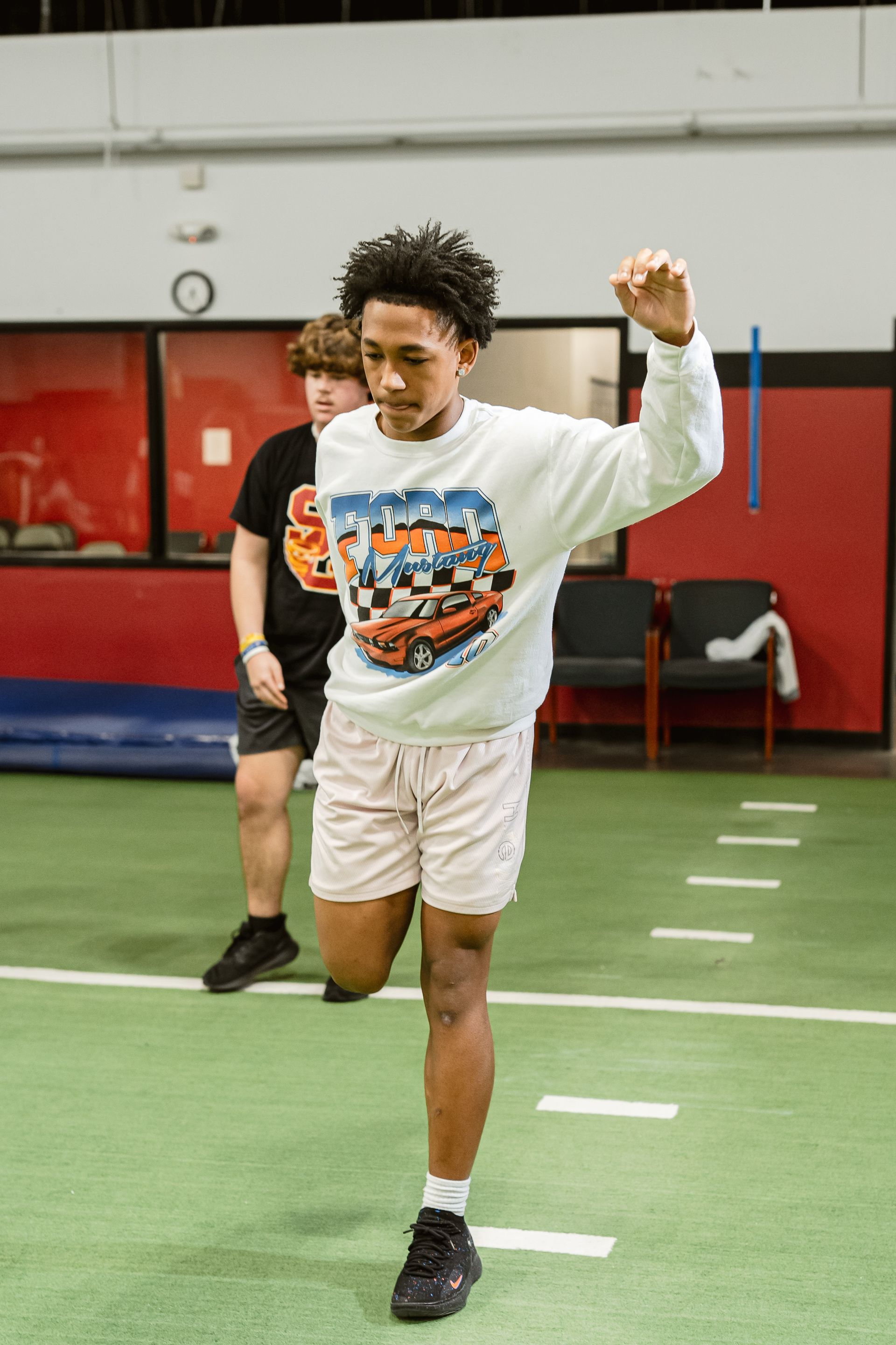 Child balancing on one leg in an indoor gym, wearing a white sweatshirt and shorts on green turf.