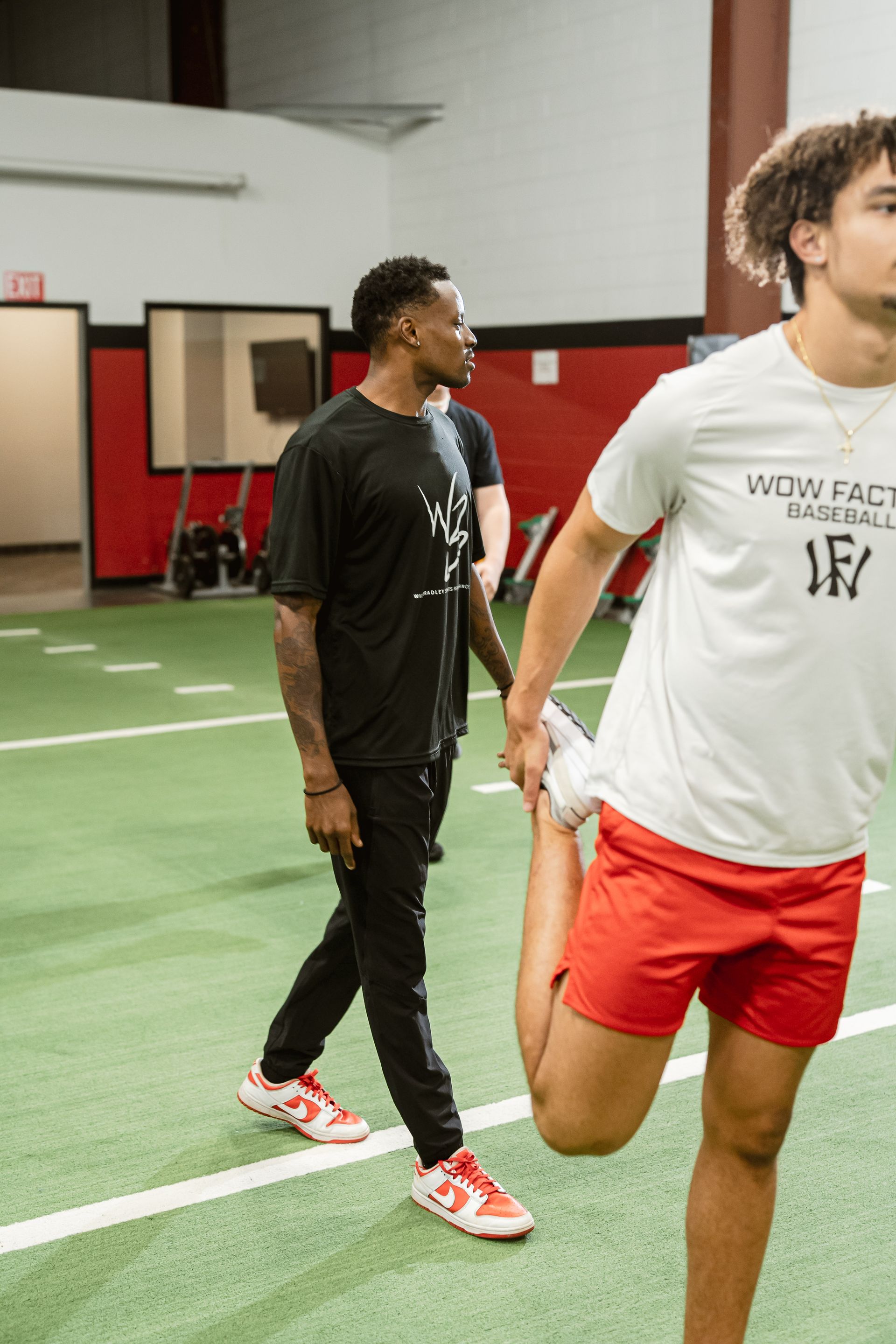 Two athletes standing on an indoor turf field, one in black and one in white and red shorts