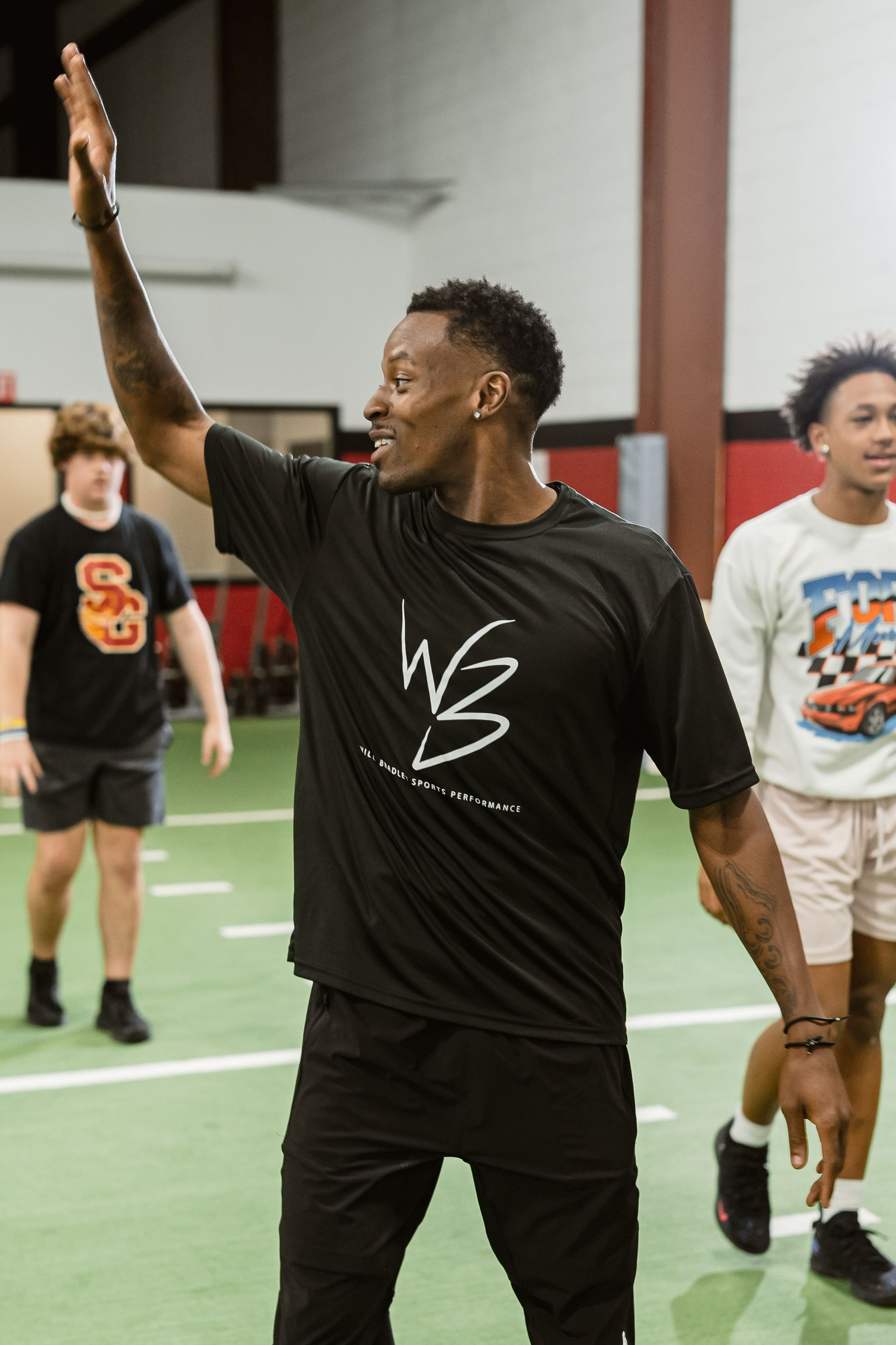 Man in black athletic wear raises his arm on an indoor court, with two people behind him.