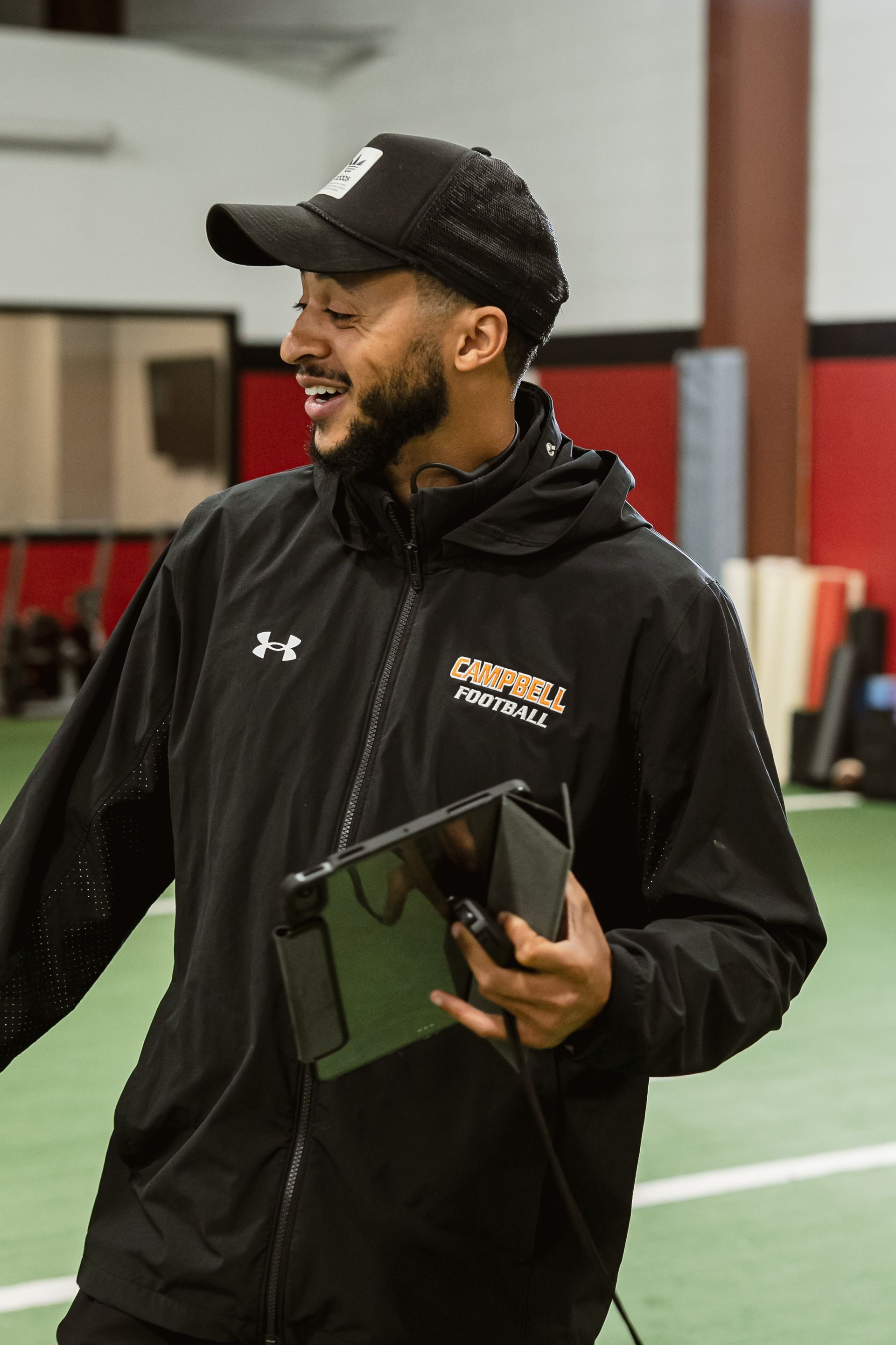 Man in black cap and jacket holding a tablet, smiling in a gymnasium.
