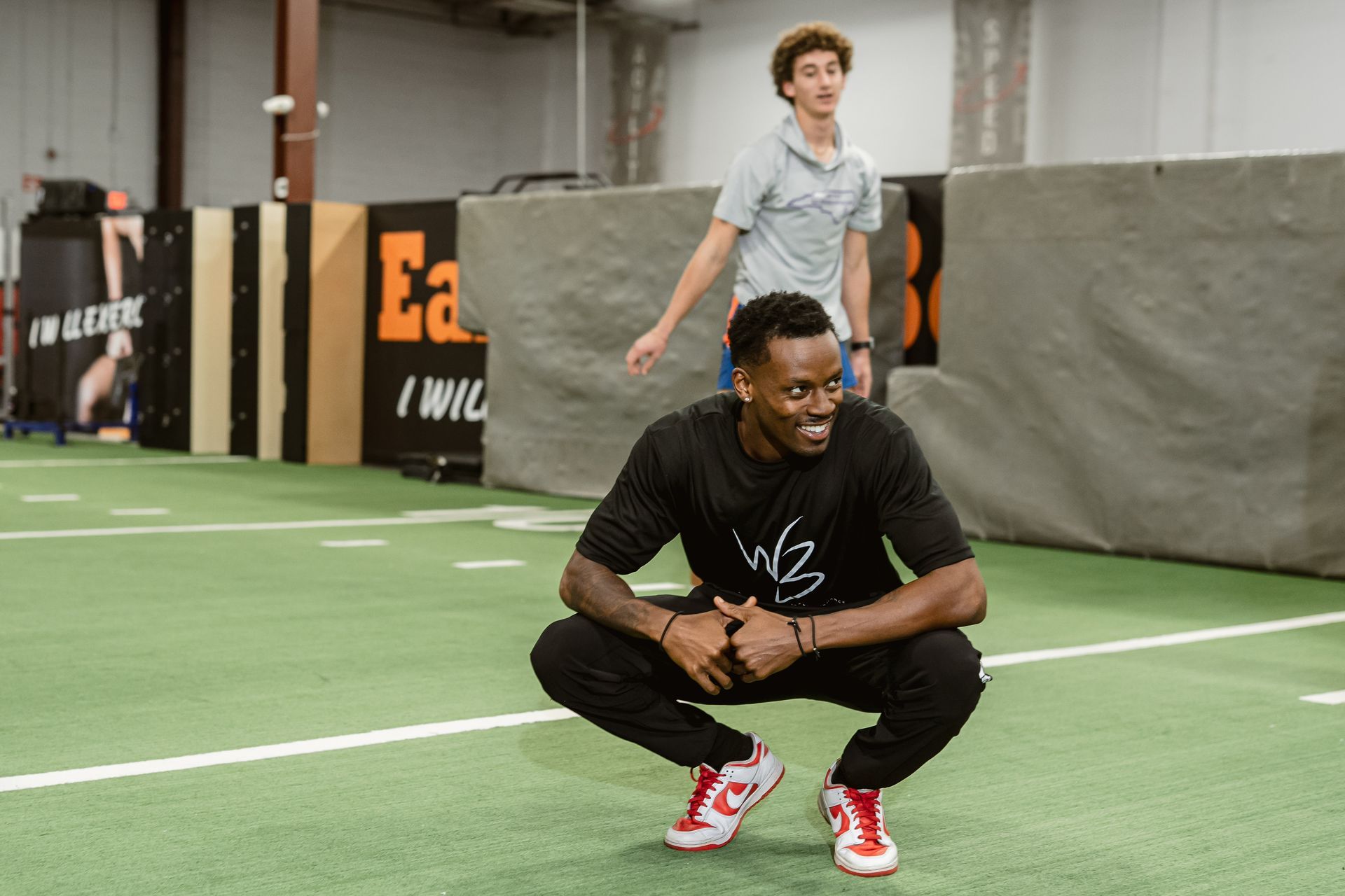 Two athletes on an indoor turf field, one crouching in front while another stands behind.