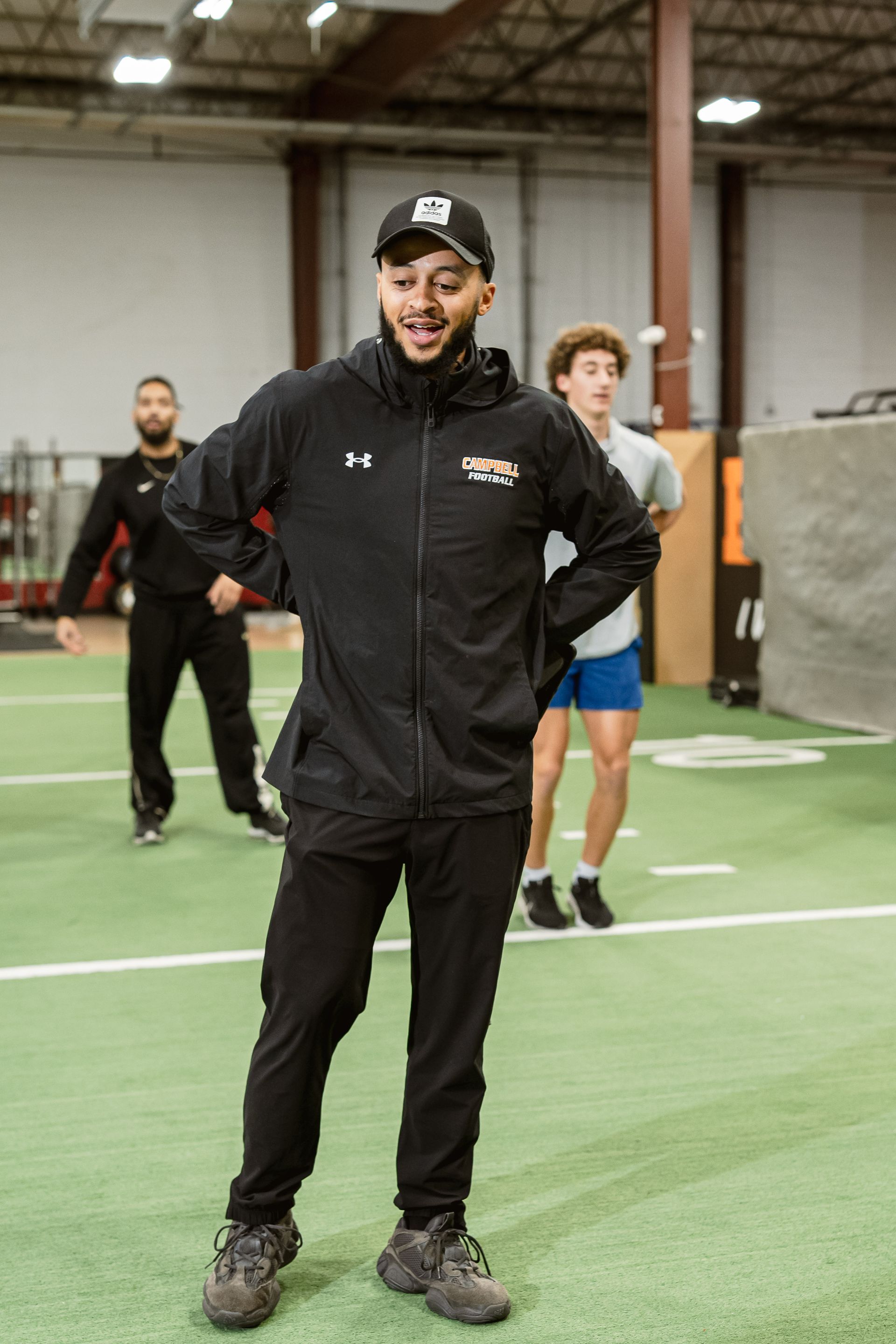 Man standing on indoor turf field, wearing a black jacket and cap, with two people blurred behind him.