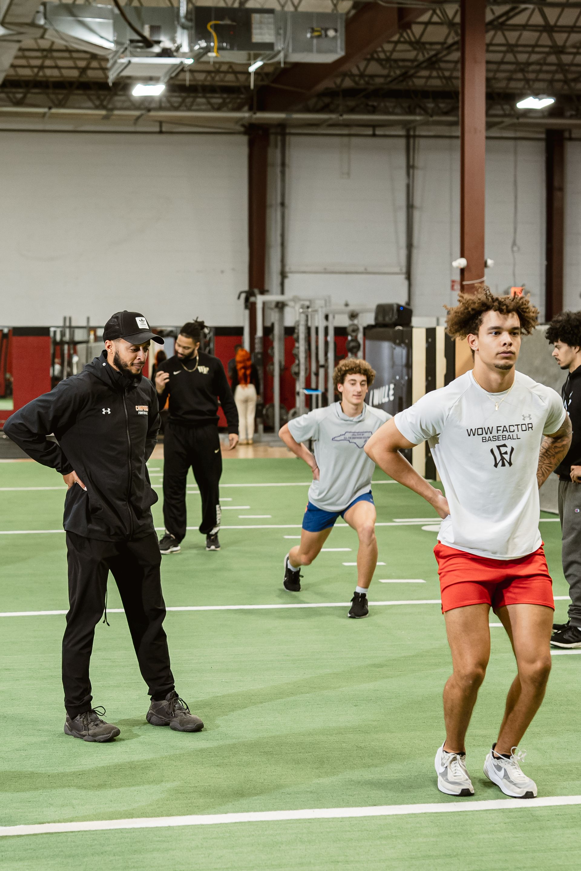 People exercising indoors on a turf field, with one man in a white shirt and red shorts in the foreground.