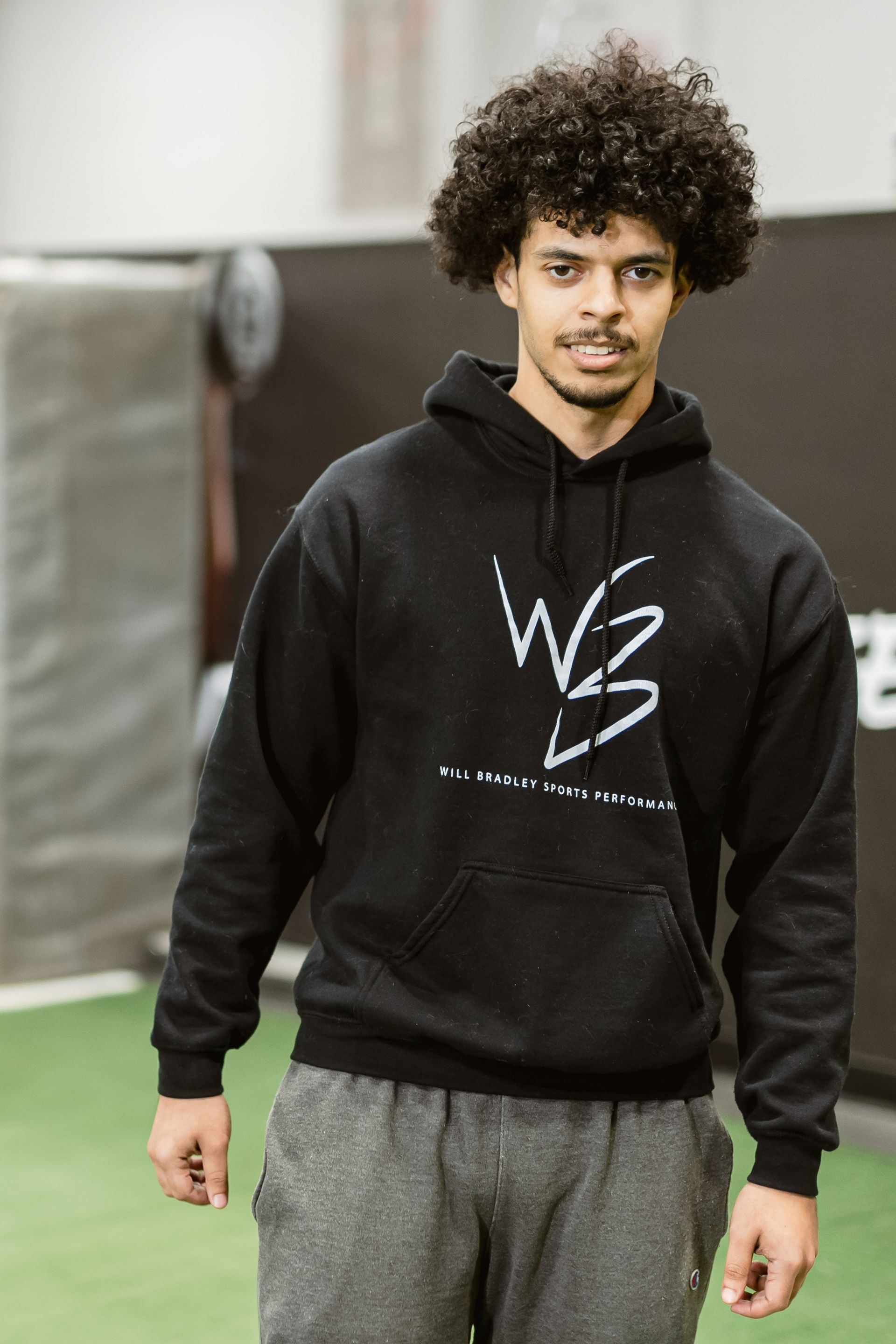 Person in a black hoodie and gray sweatpants standing indoors on a green floor.