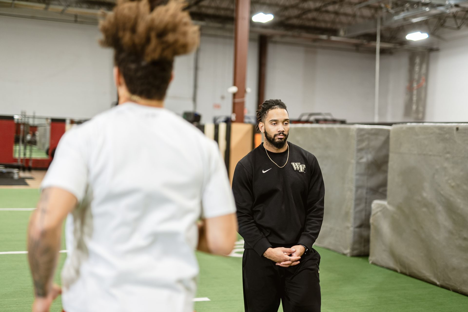 Two people on an indoor turf field, one in black facing a blurred runner in white.
