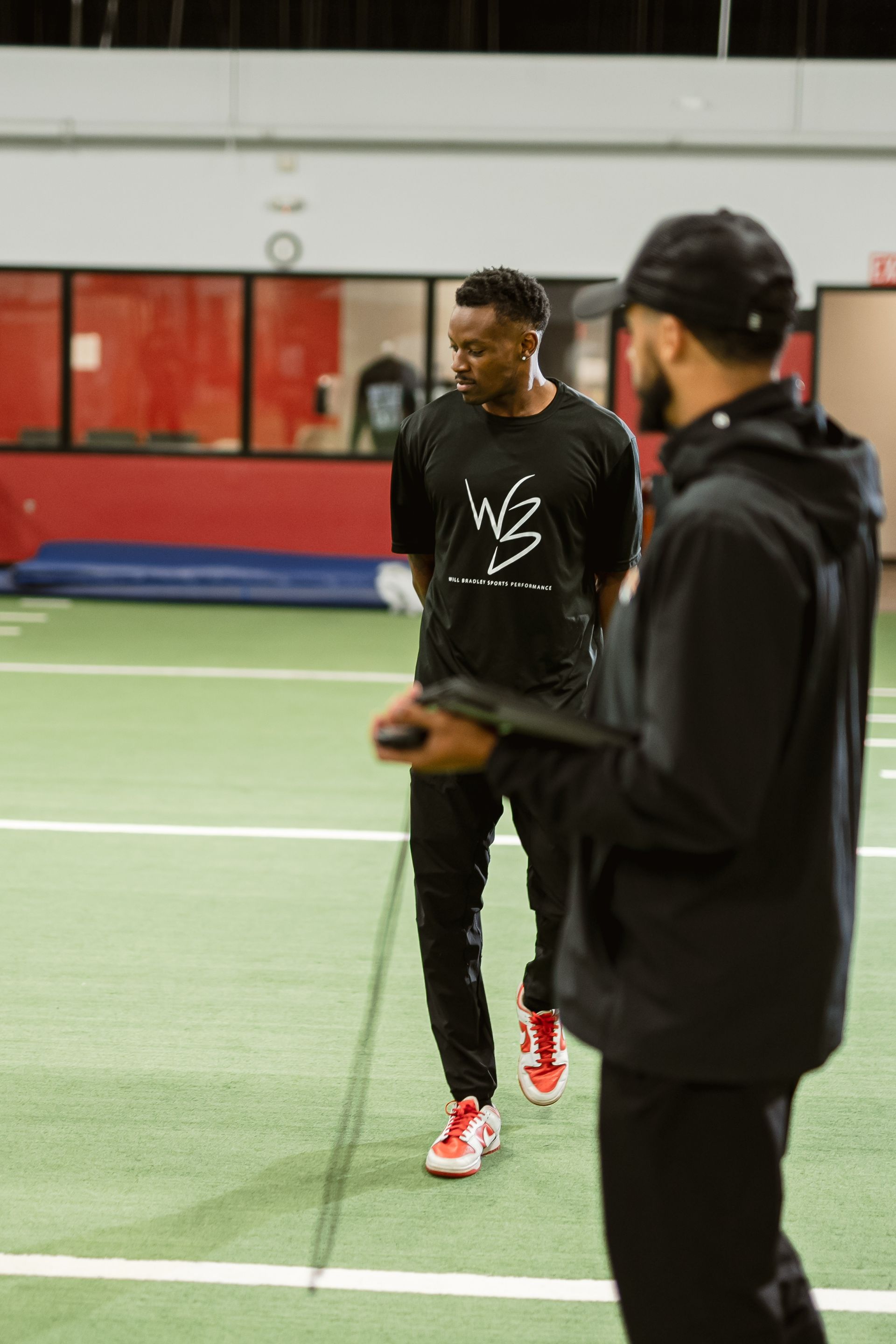 Two people on an indoor turf field, one standing in black and red sneakers holding a ball.
