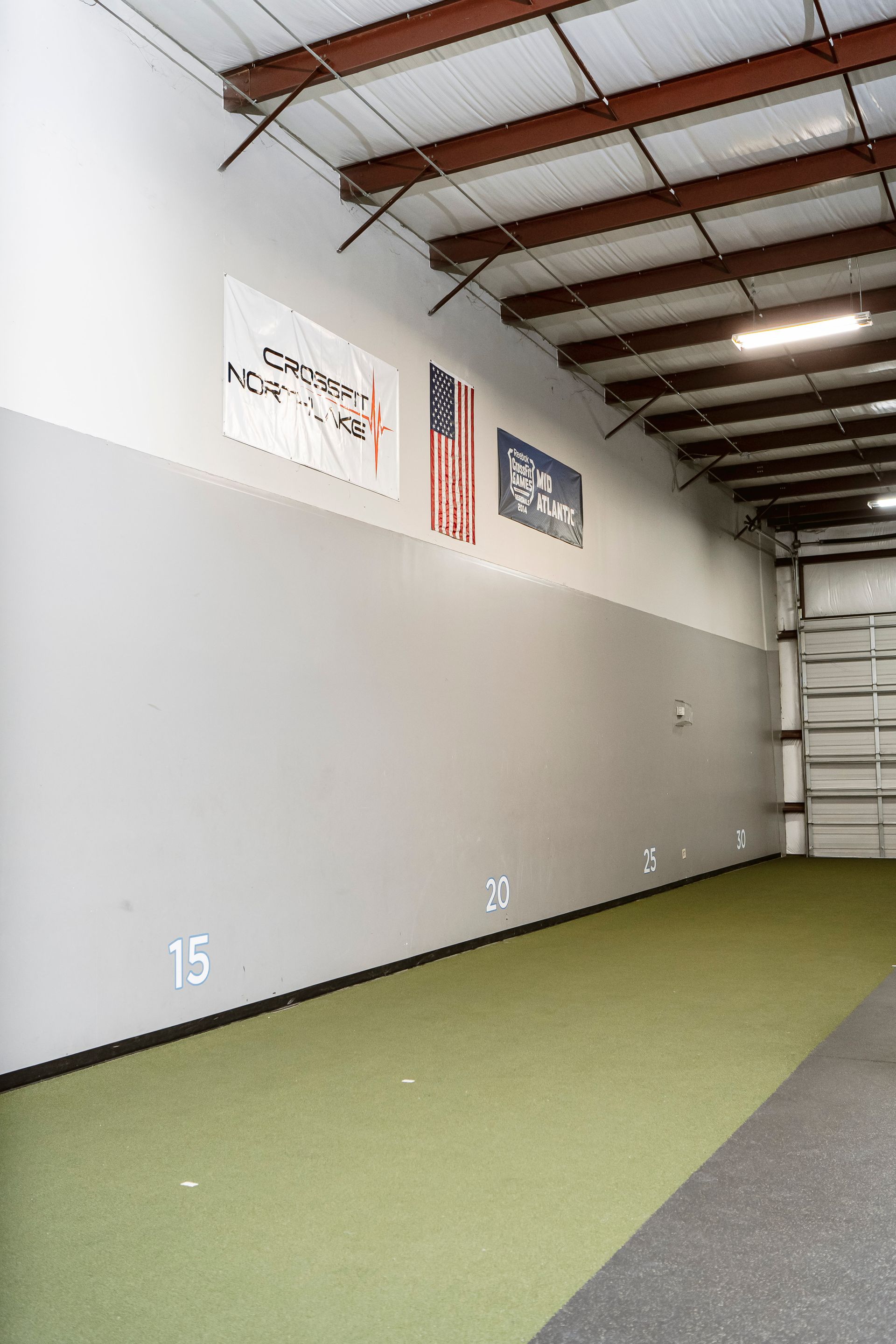 Empty warehouse gym with turf lane, gray wall, overhead beams, and signs near the ceiling