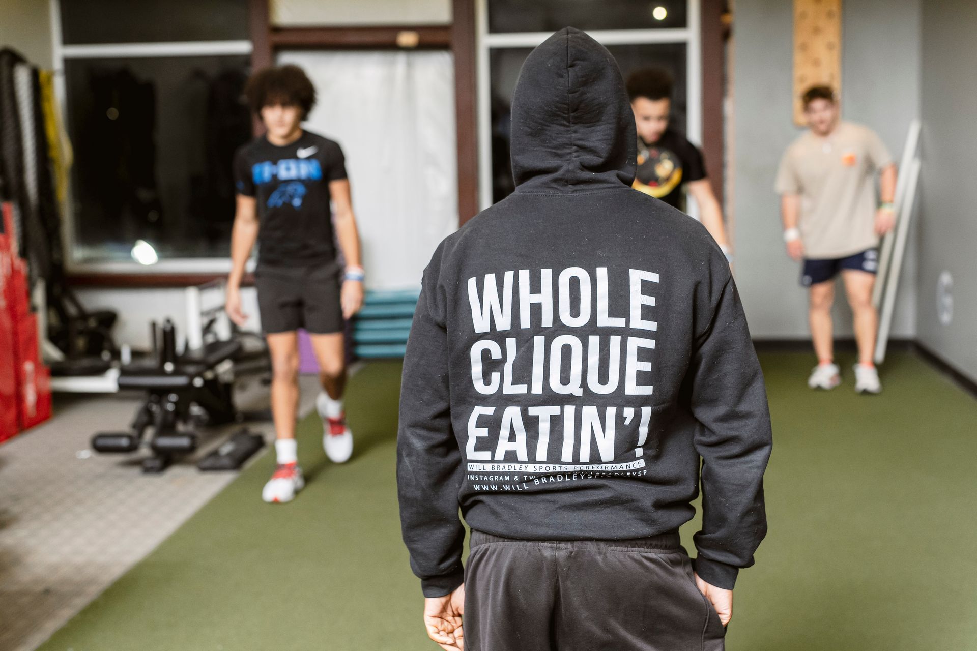 Person in black hoodie with “WHOLE CLIQUE EATIN!” stands in a gym facing two athletes.