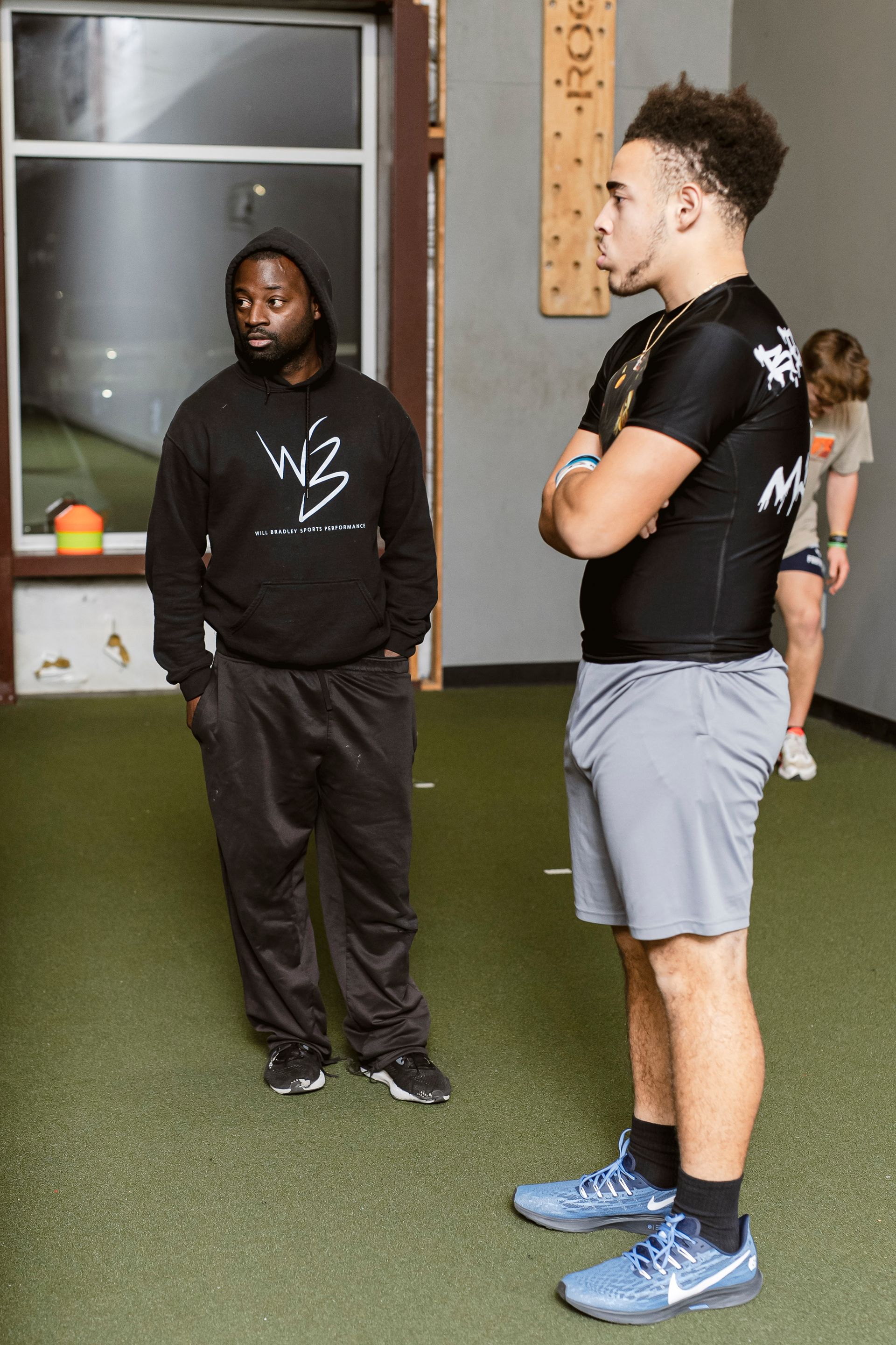 Three people standing in an indoor gym, two in the foreground facing each other.
