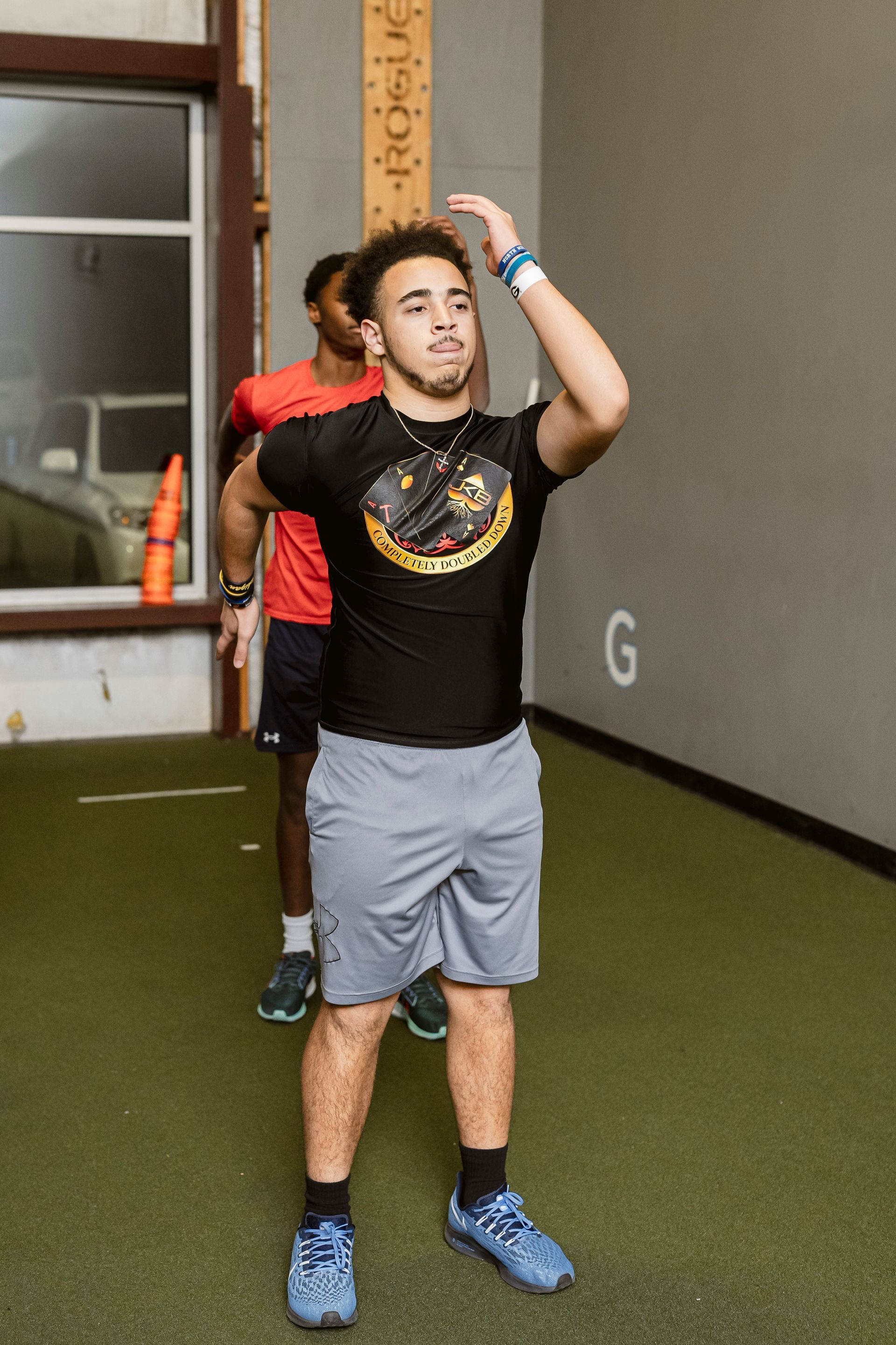 Person standing indoors with arm raised, wearing a black shirt, gray shorts, and blue shoes.