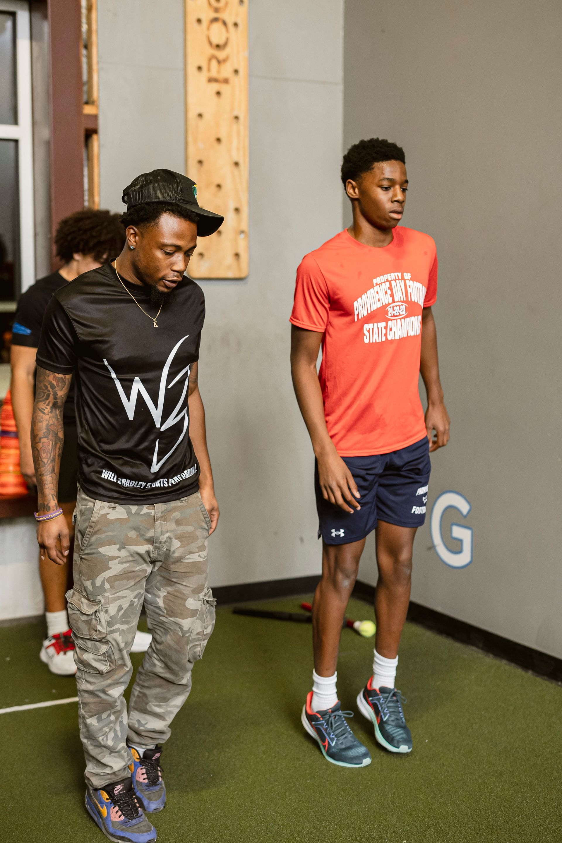 Two people standing indoors on green turf, one looking down and one facing forward, near a gray wall