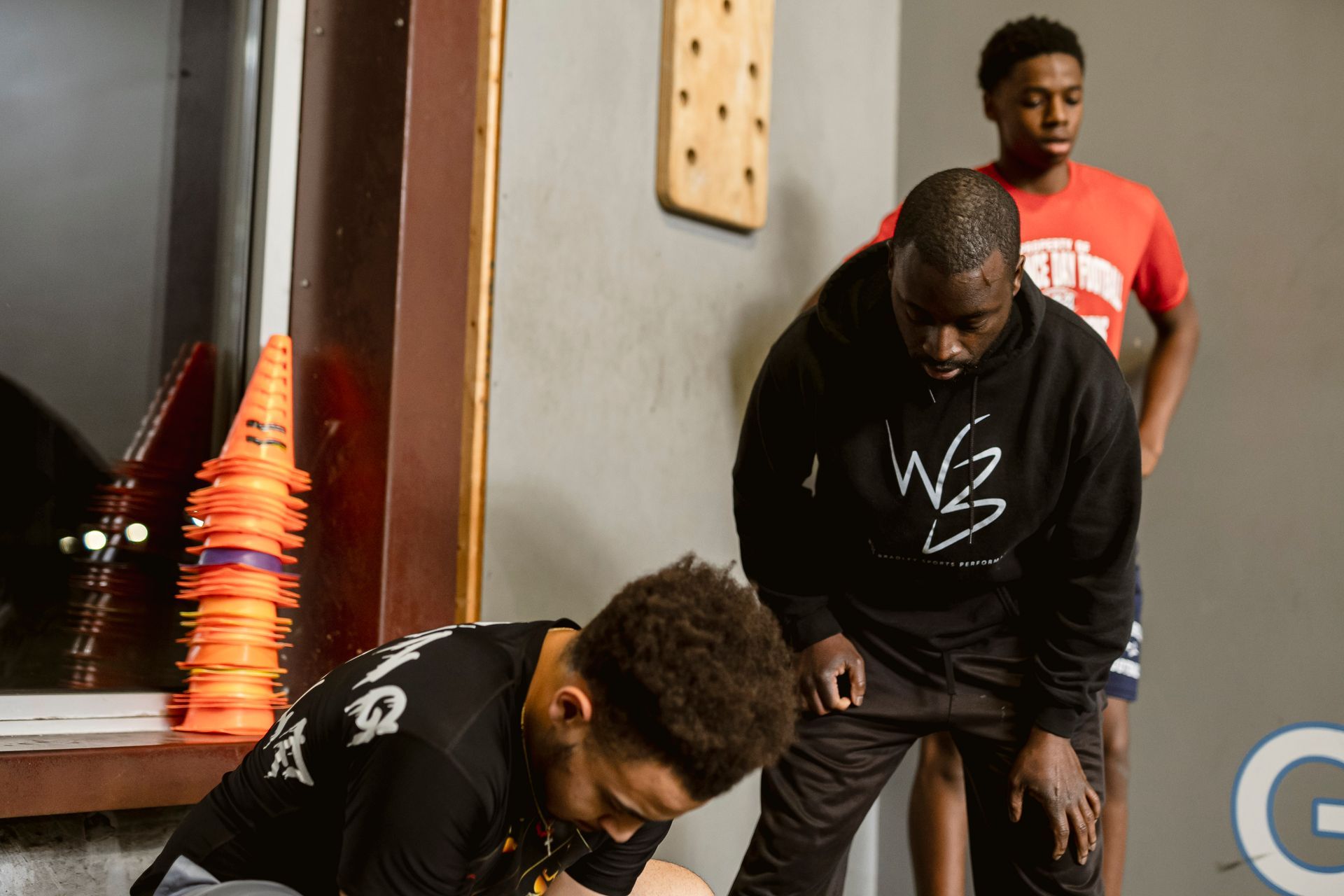 Three people in athletic gear stretching indoors near cones and a doorway.