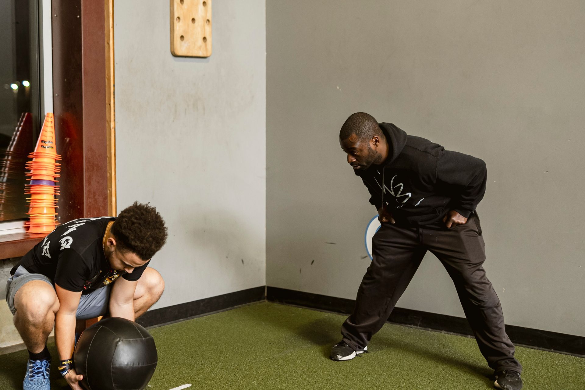 Two people in a gym stretching near a black medicine ball on turf flooring.