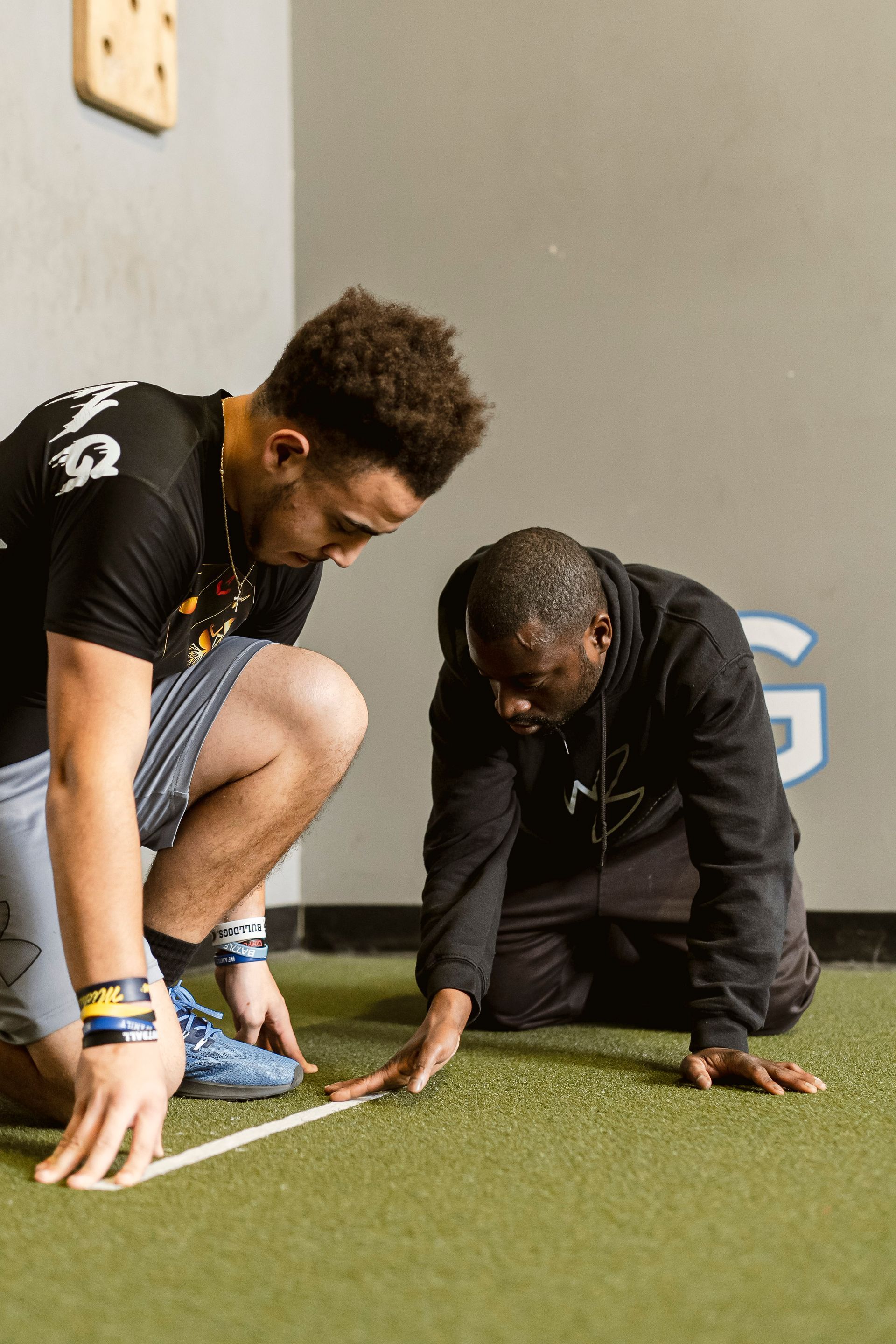 Two athletes kneel on turf, focused on a starting line in an indoor training area.