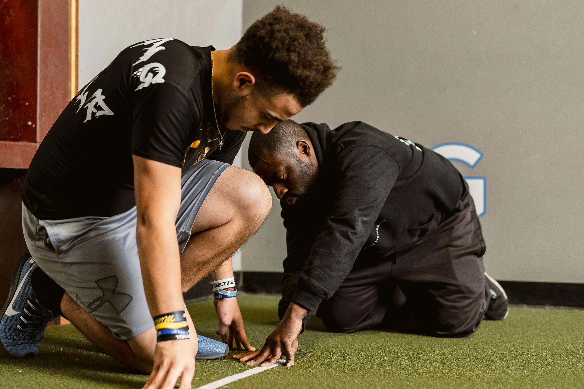 Two people kneel on turf, measuring or marking a white line in a sports facility.