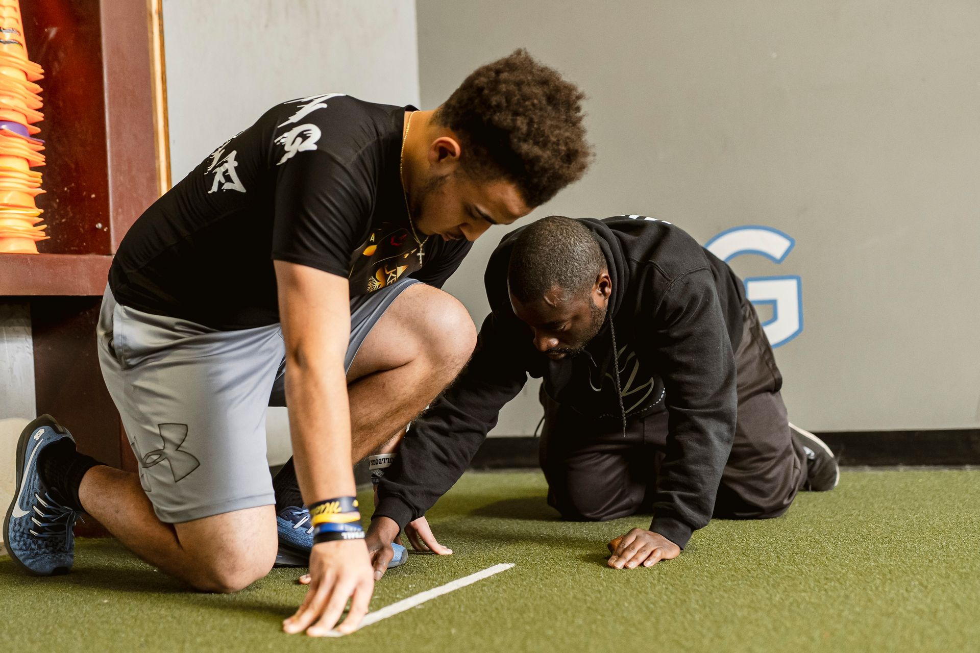 Two athletes kneel on turf, practicing a ground drill in an indoor training facility.