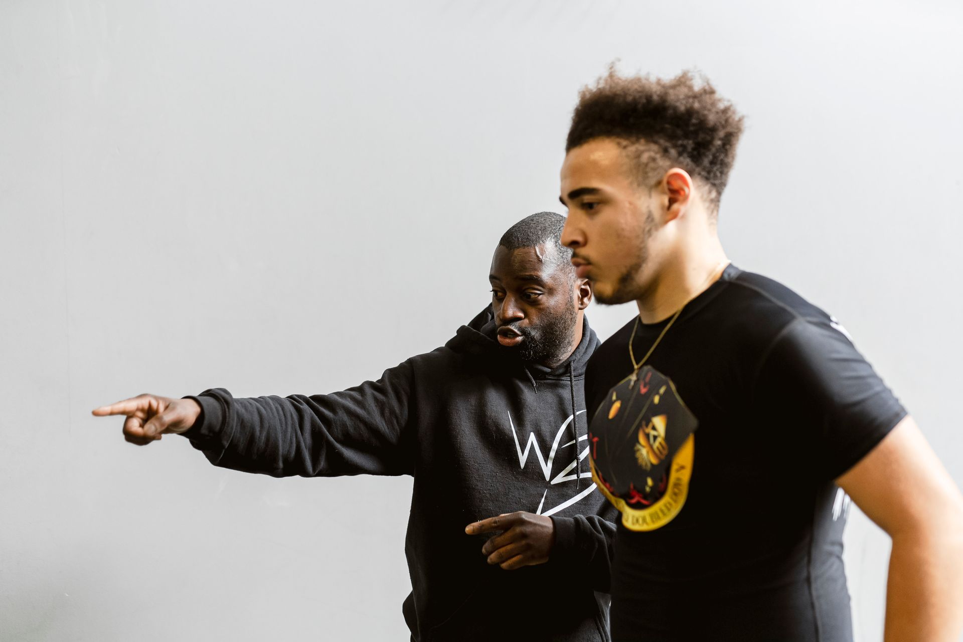 Two men in black shirts indoors, one pointing left while the other looks on.