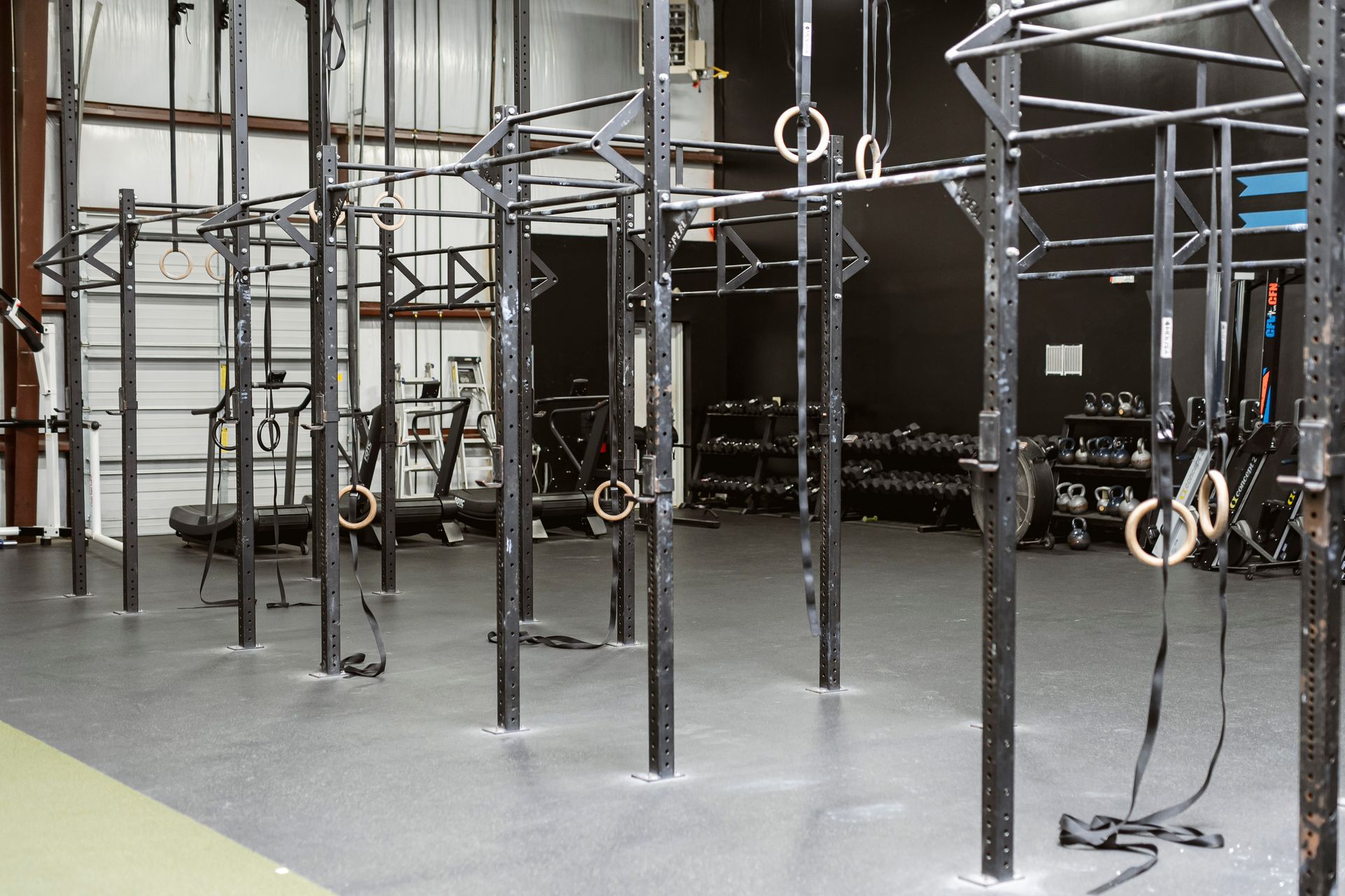Indoor gym with black metal climbing rig and rings on a gray floor, plus a bicycle in the back.