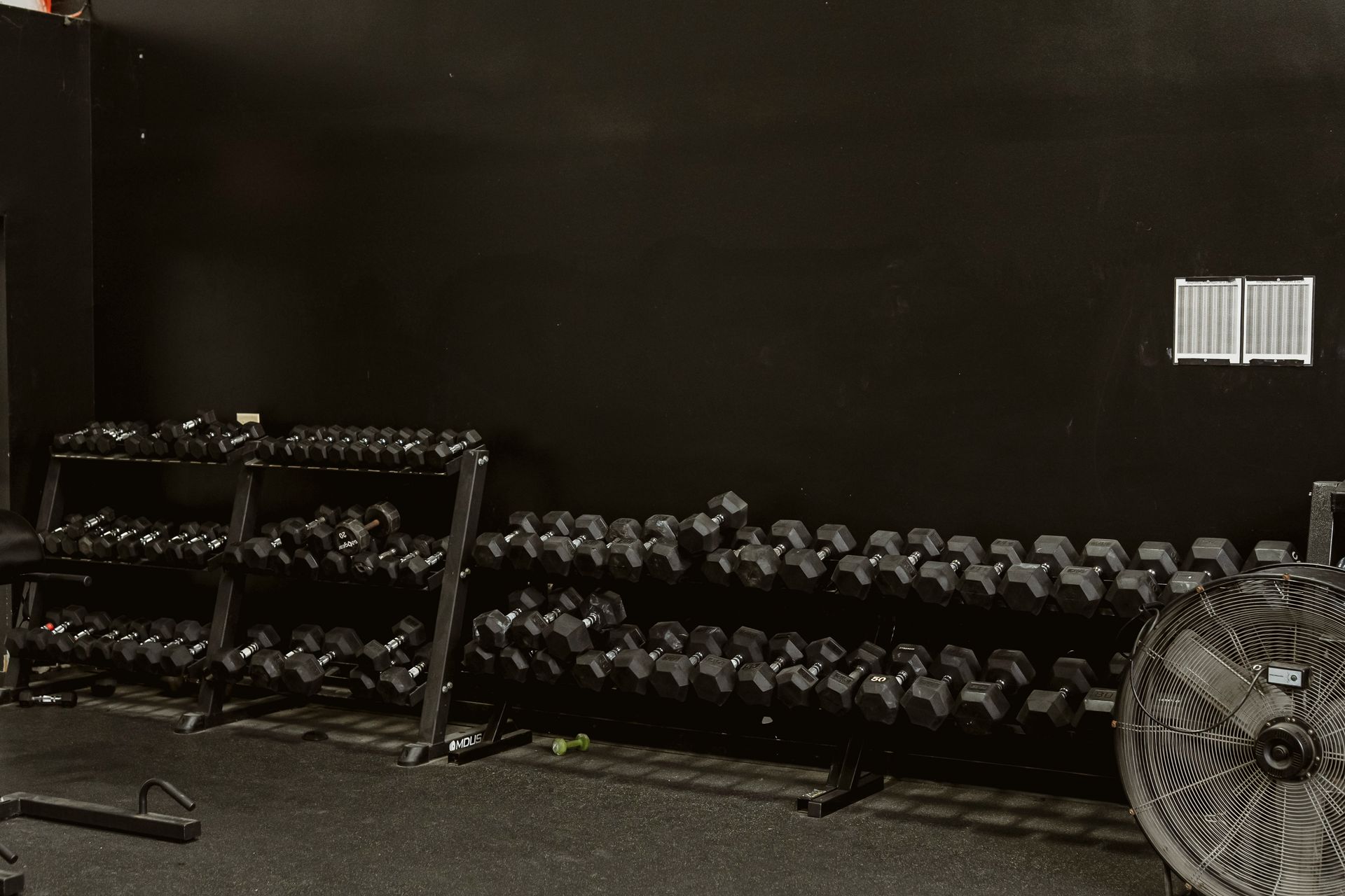 Gym with rows of dumbbells, a rack, and a large fan against a dark wall