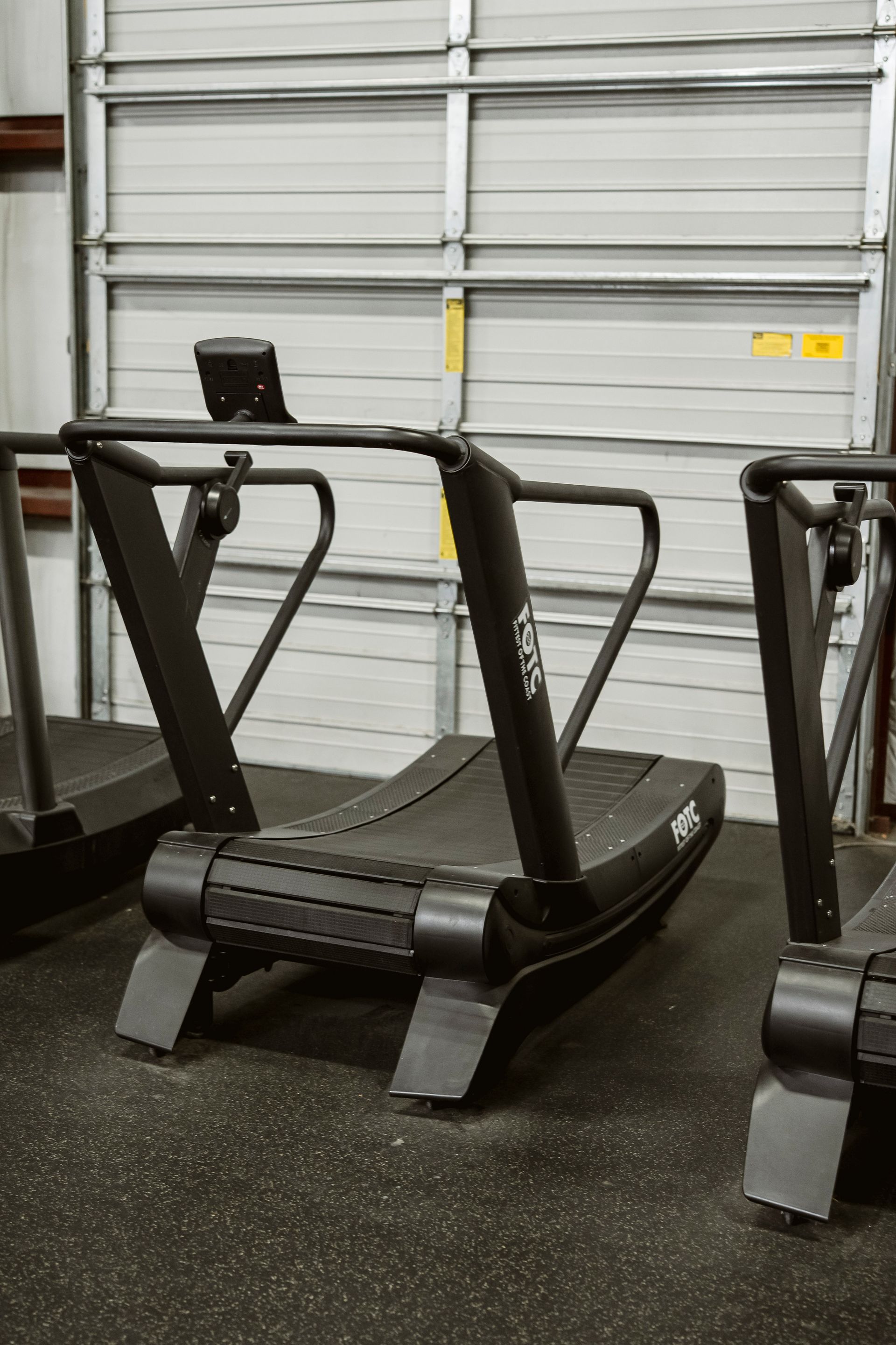 Treadmill exercise machine in a gym, black with silver frame in front of a gray garage door.